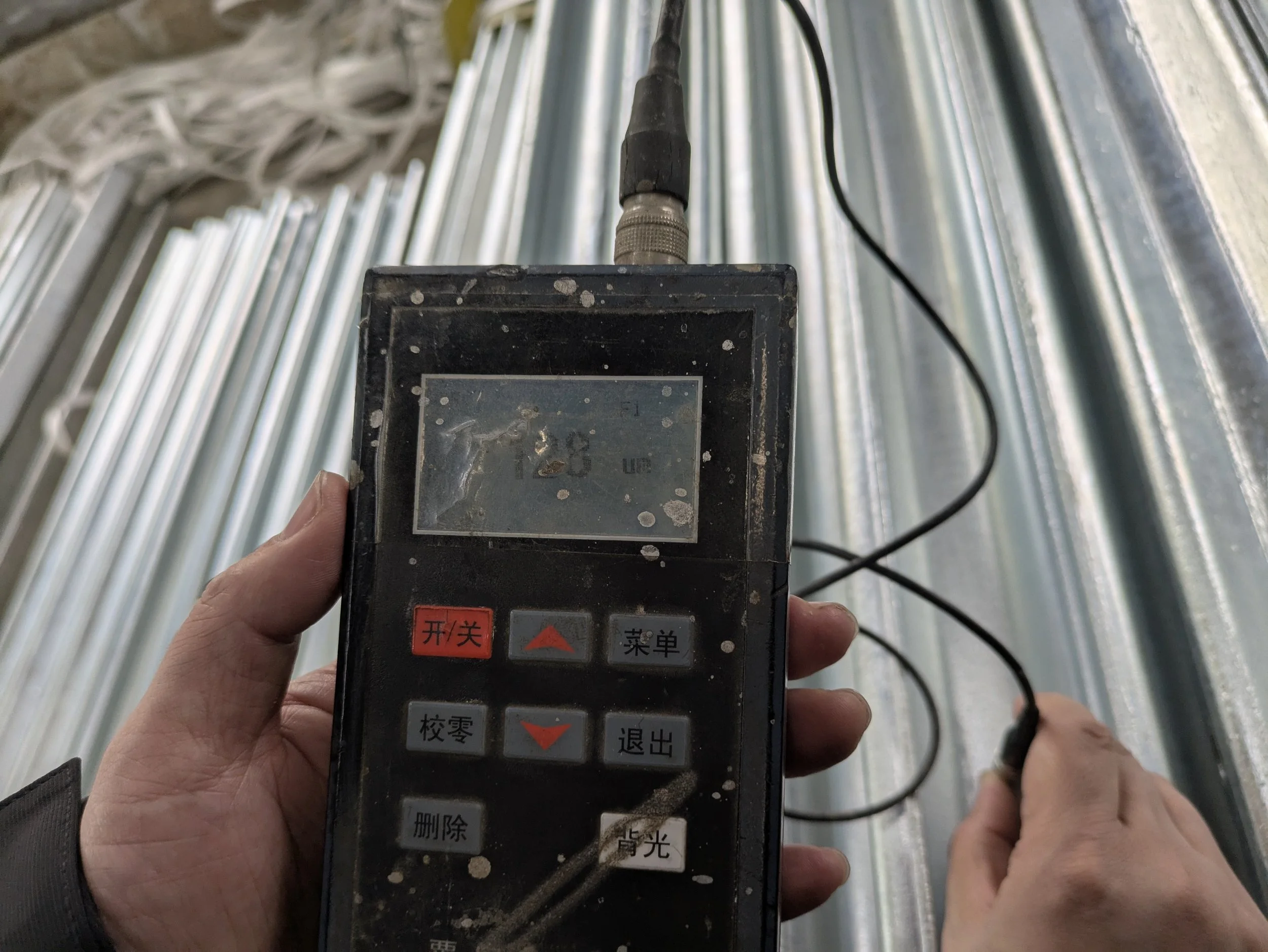 A person holding a digital instrument with Chinese characters and red and gray buttons, measuring temperature or electrical parameters in a metal structure or pipes.