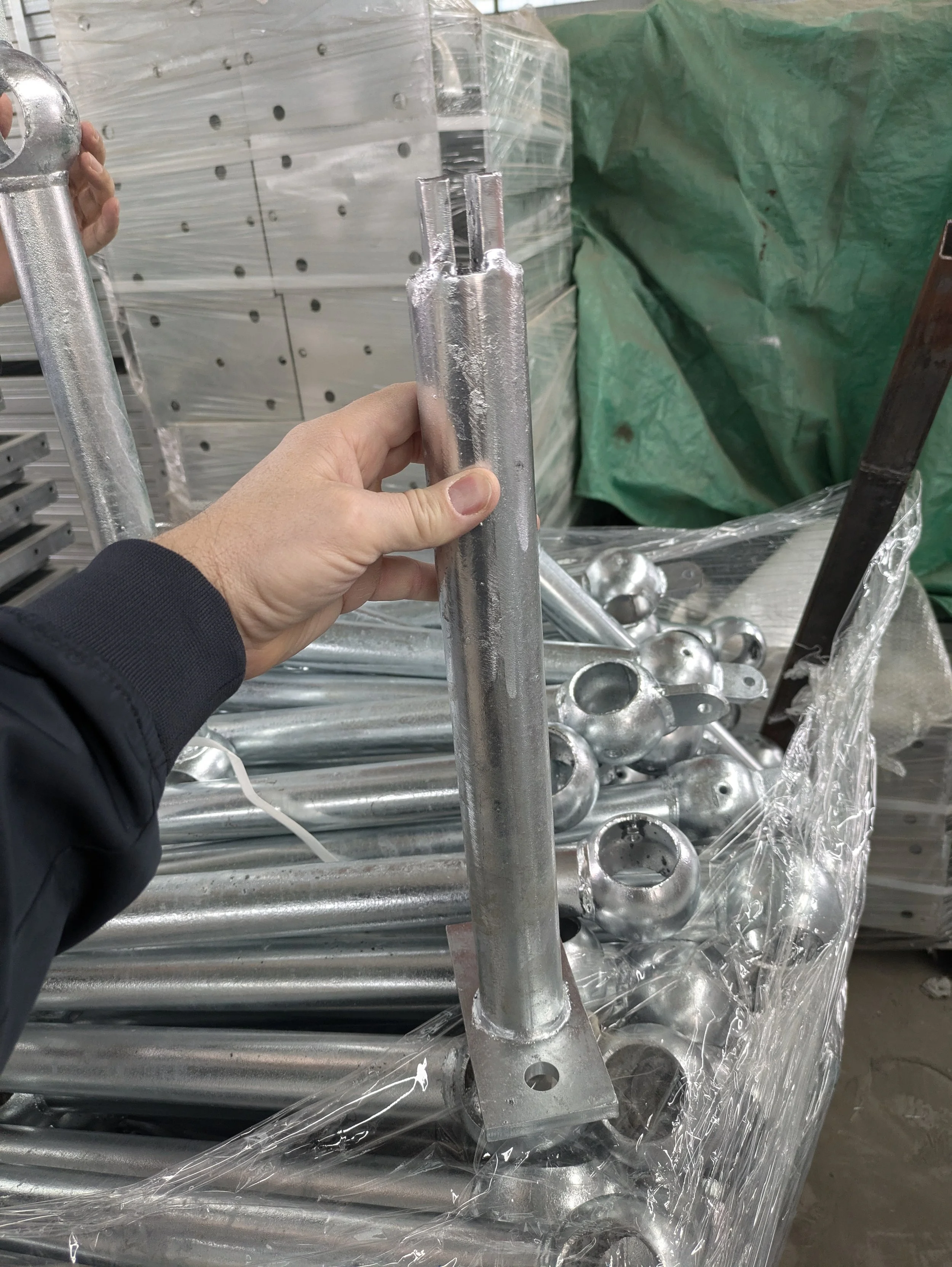 A person holding a metal pole with a U-shaped attachment, surrounded by other metal parts and fittings on a pallet in a warehouse or manufacturing setting.