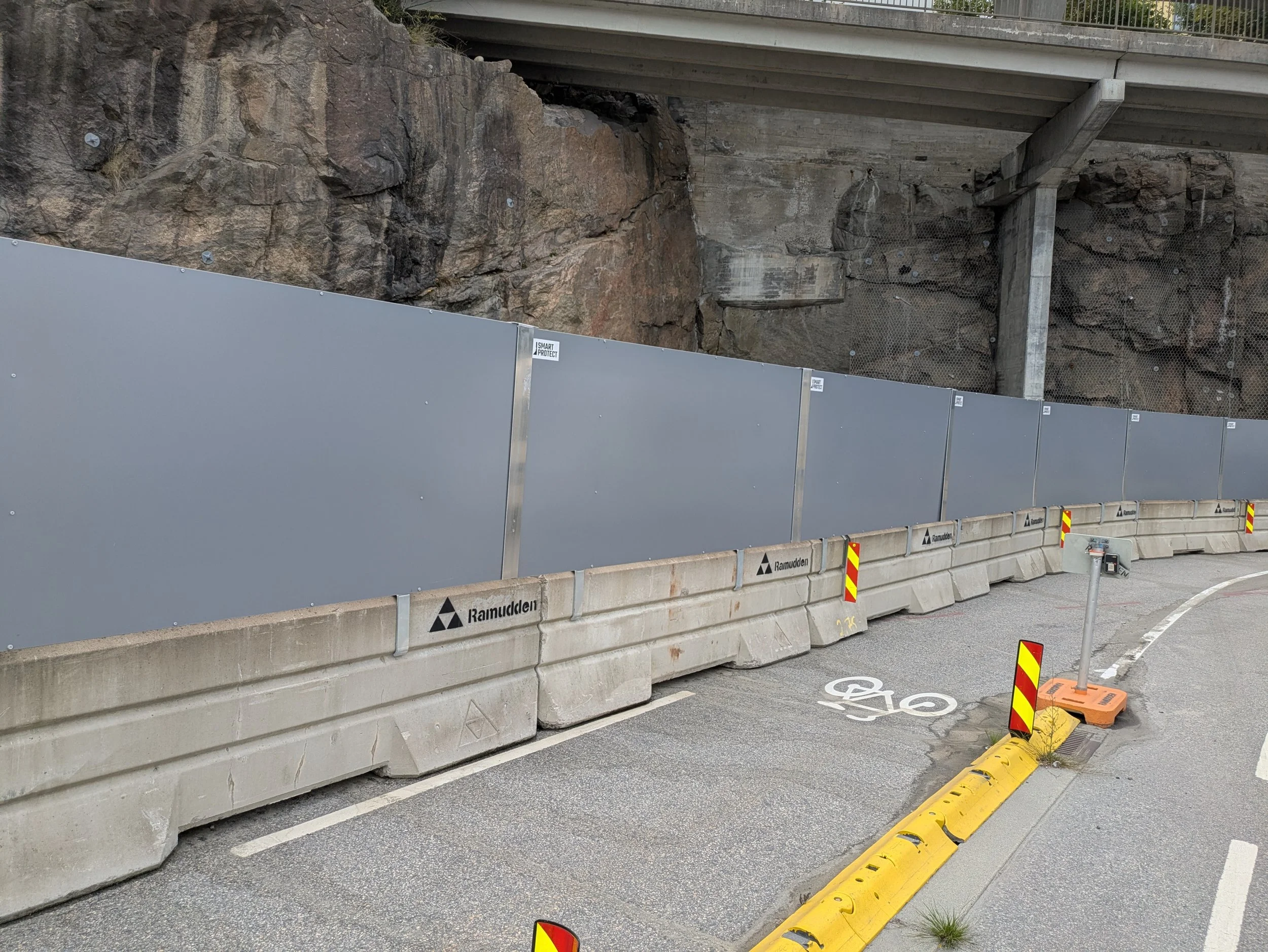 Construction barrier with gray panels and yellow traffic delineator on a bicycle lane next to rocky terrain and an overpass.