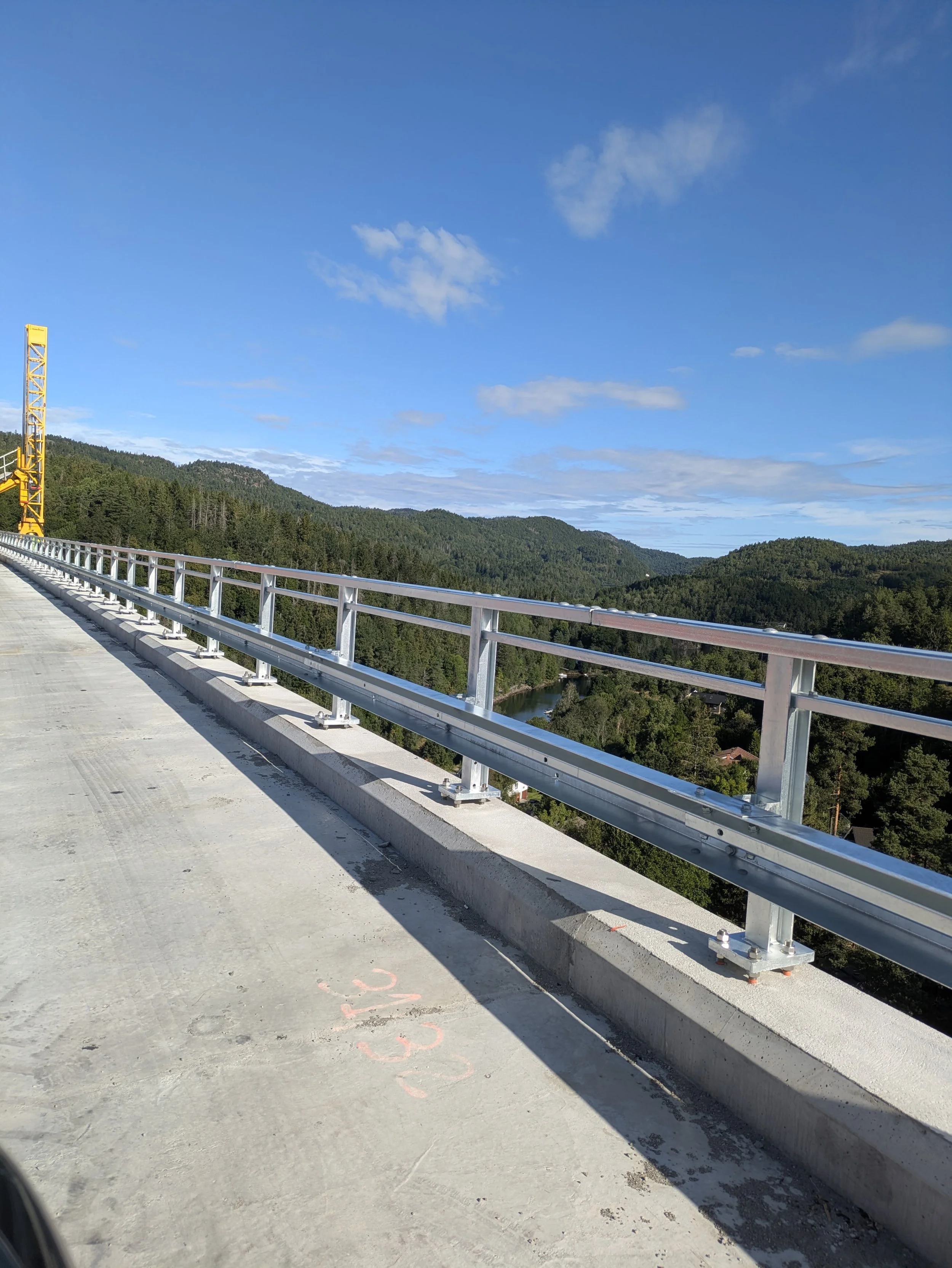 Steel railing on a concrete highway bridge with a scenic view of forested hills and a river below under a partly cloudy sky.