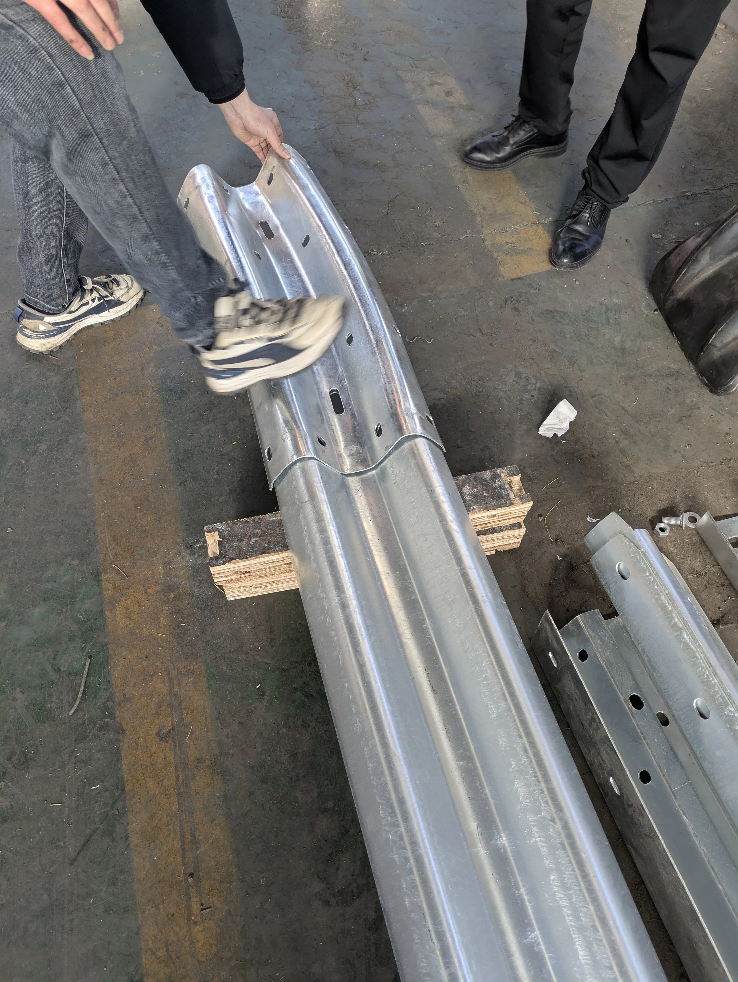 Two people working with a metal bumper at a workshop, one wearing gray jeans and sneakers, the other in black pants and shoes, with the bumper resting on wooden blocks on the concrete floor.