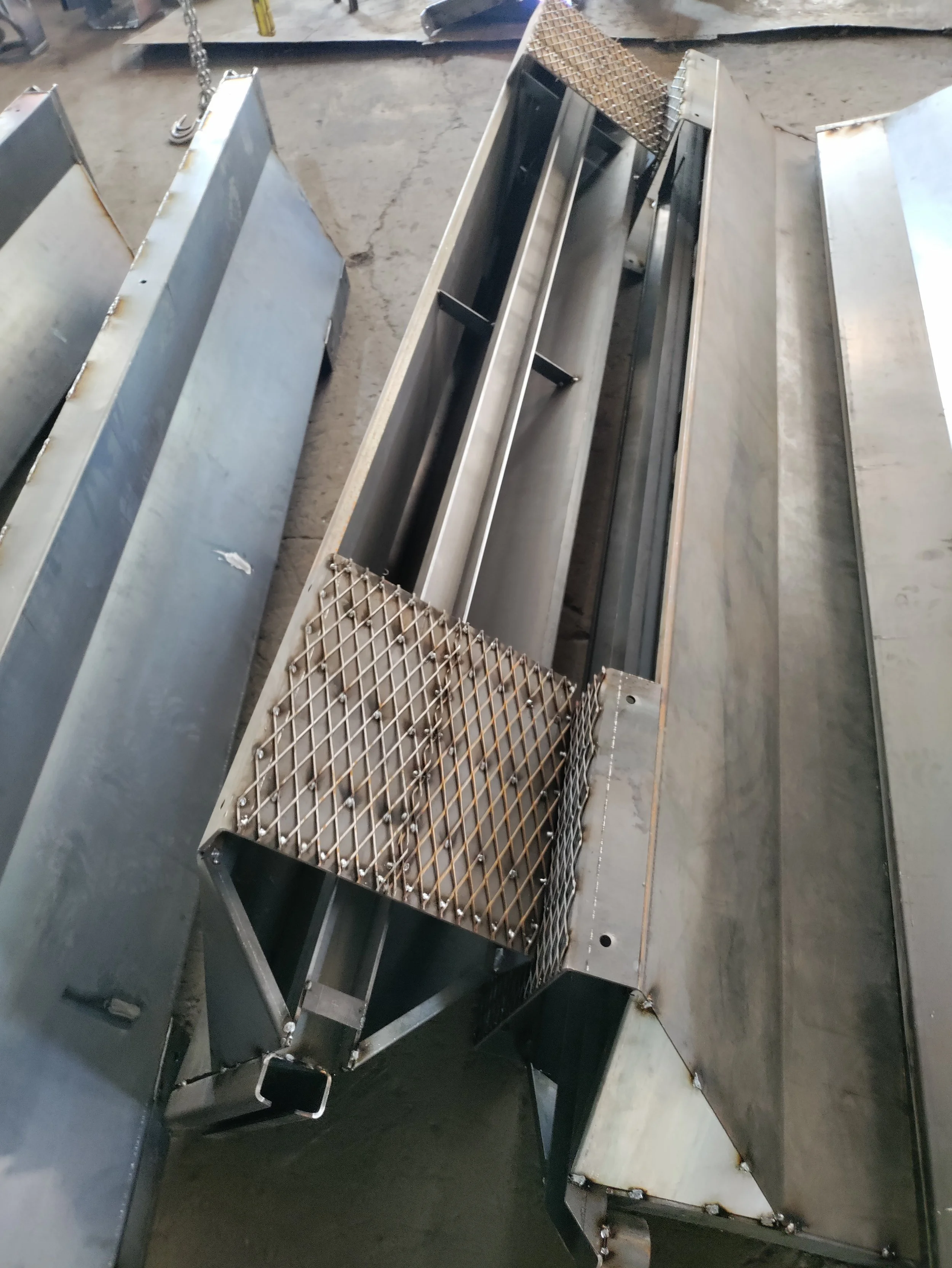 Large stainless steel industrial equipment parts, including a metal grate and framework, placed on a concrete floor in an industrial setting.