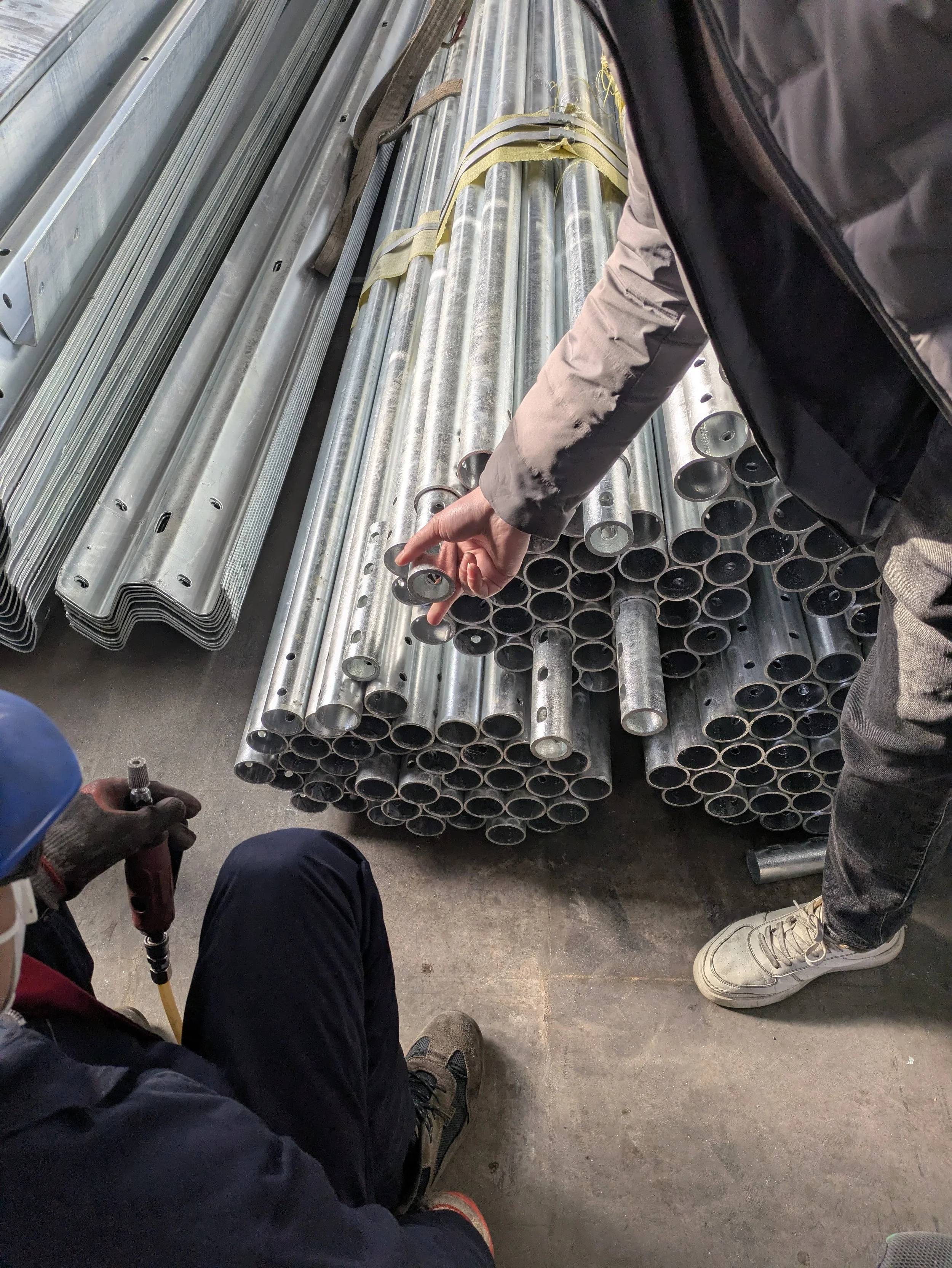 Two workers examining metal pipes in a warehouse or factory setting, with one worker holding a bolt and the other pointing at a pipe.