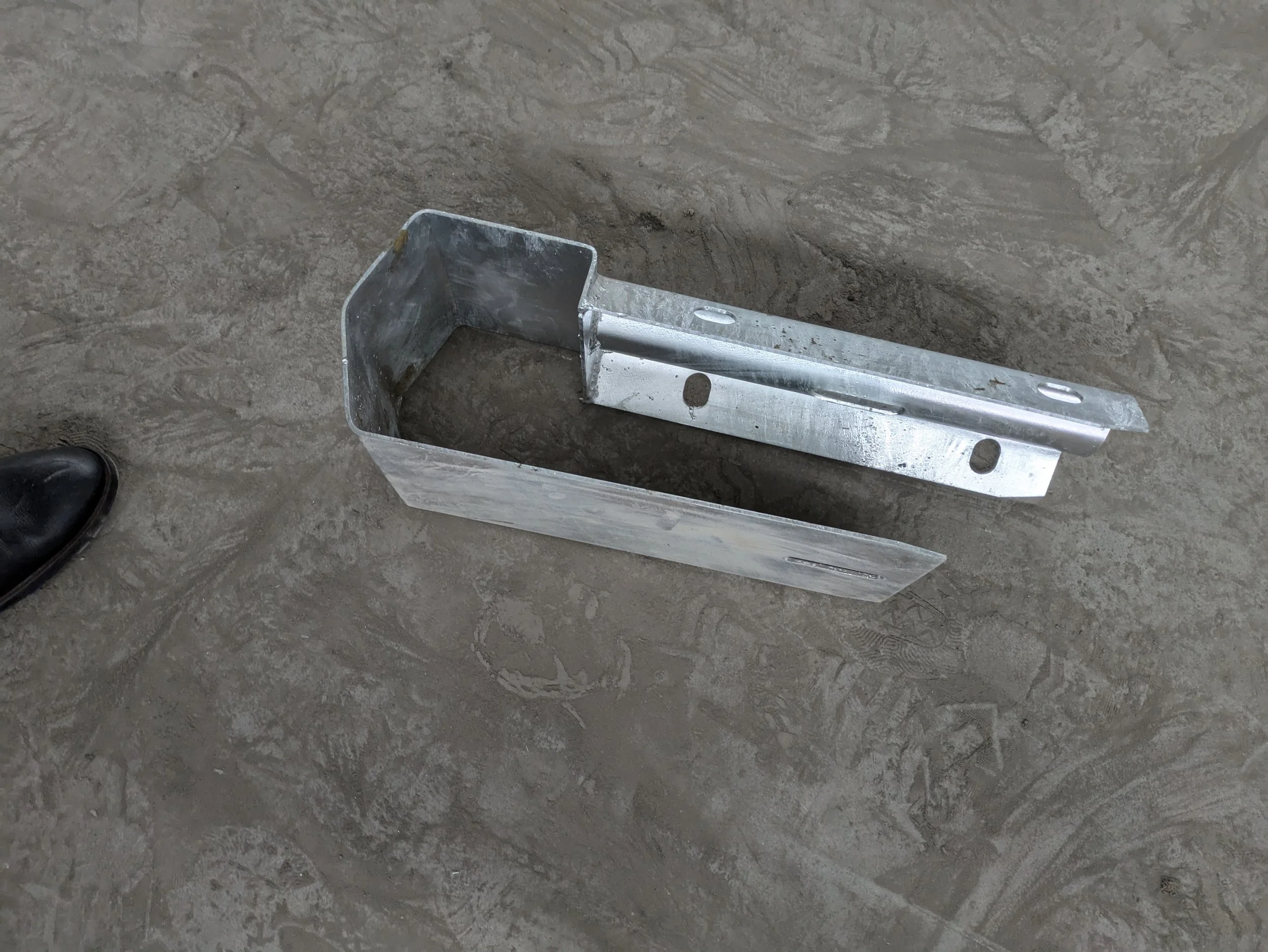 Metal bracket lying on a dusty concrete floor, partially visible shoe of a person on the left side.