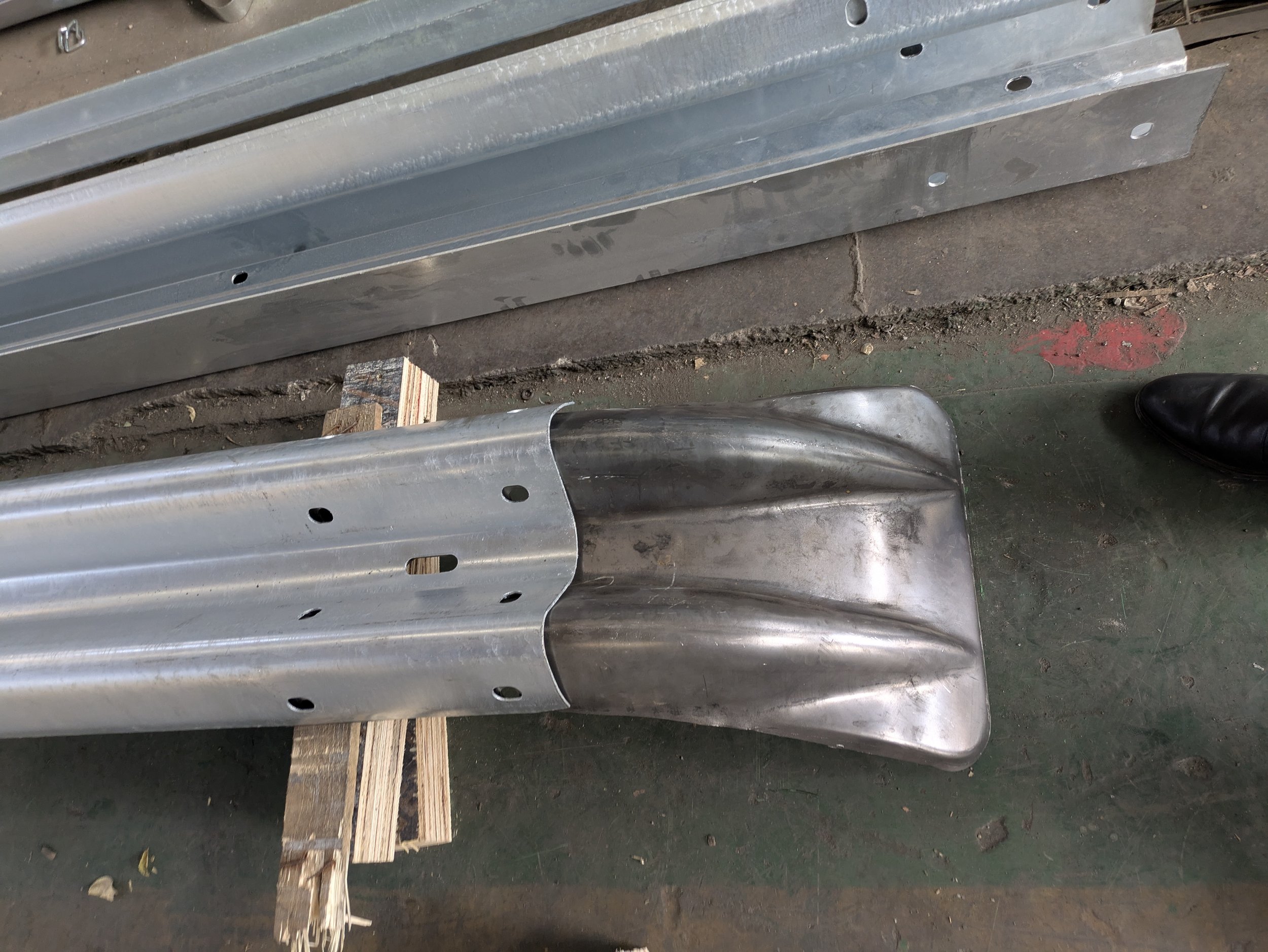 Metal car part with a curved shape and multiple holes, resting on a wooden block on a workshop floor, with additional metal pieces stacked above.