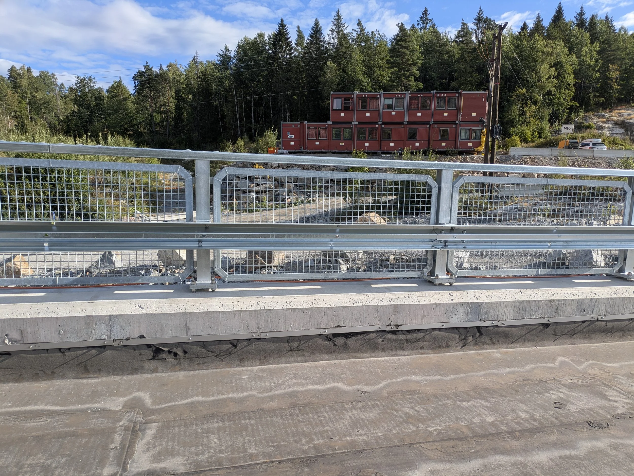 View from a bridge showing a construction site with a stack of red modular units, surrounded by trees and a road in the background.