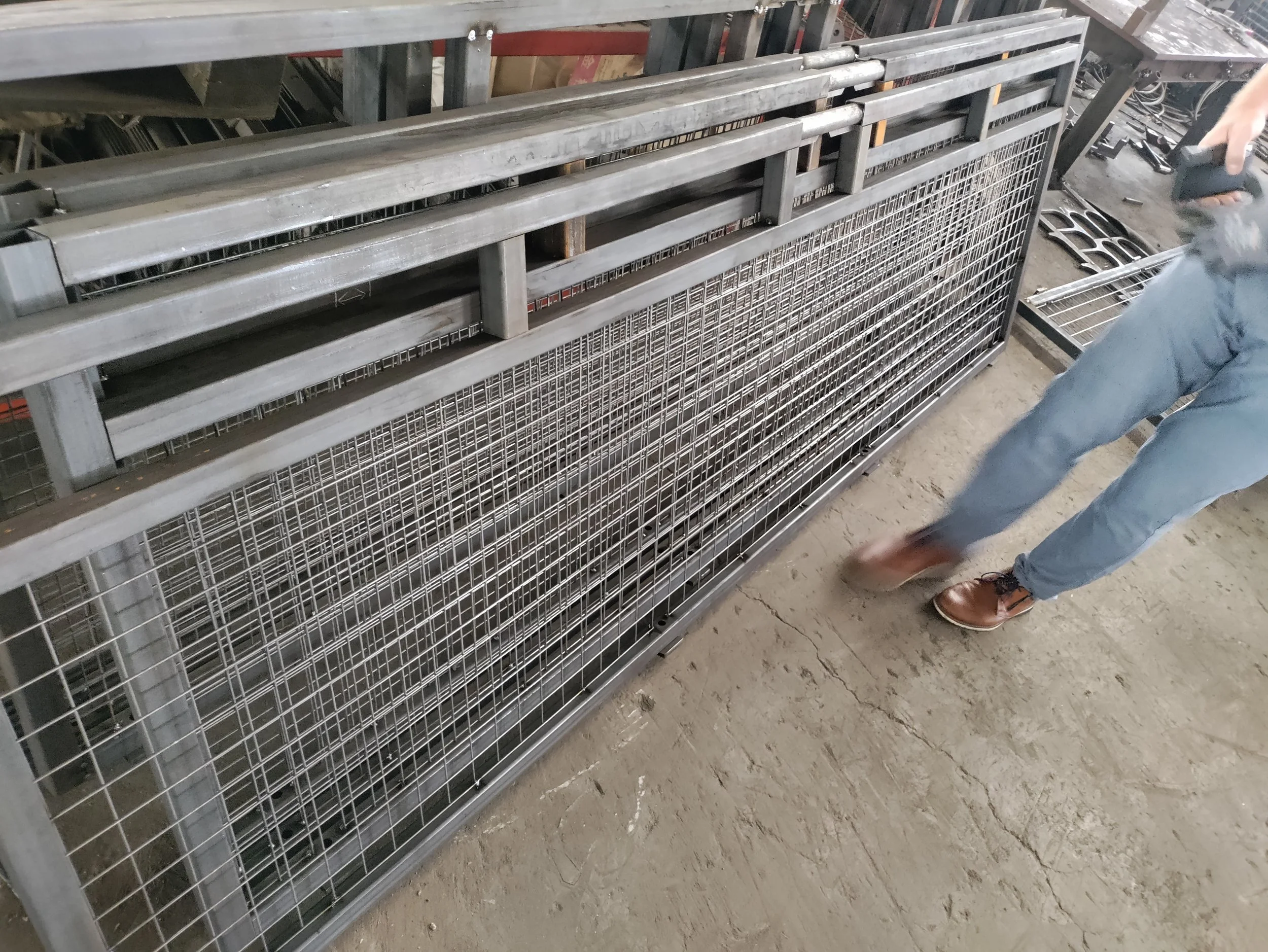 A large metallic wire storage rack with multiple shelves, located in a warehouse or workshop. Part of a person wearing jeans and brown shoes is visible on the right side of the image.