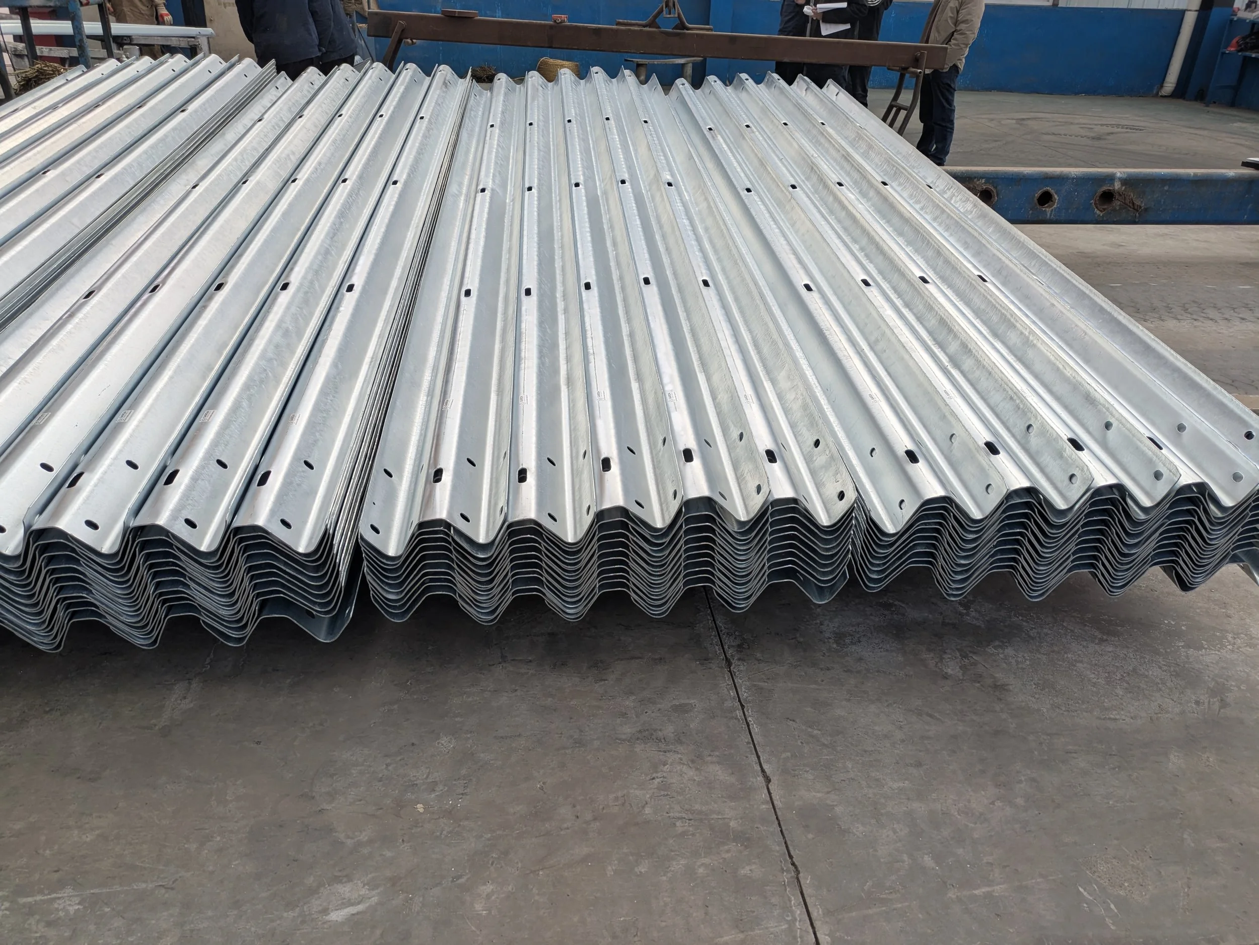 Stack of metal roof tiles with a zigzag edge and hole patterns in a factory or warehouse setting.