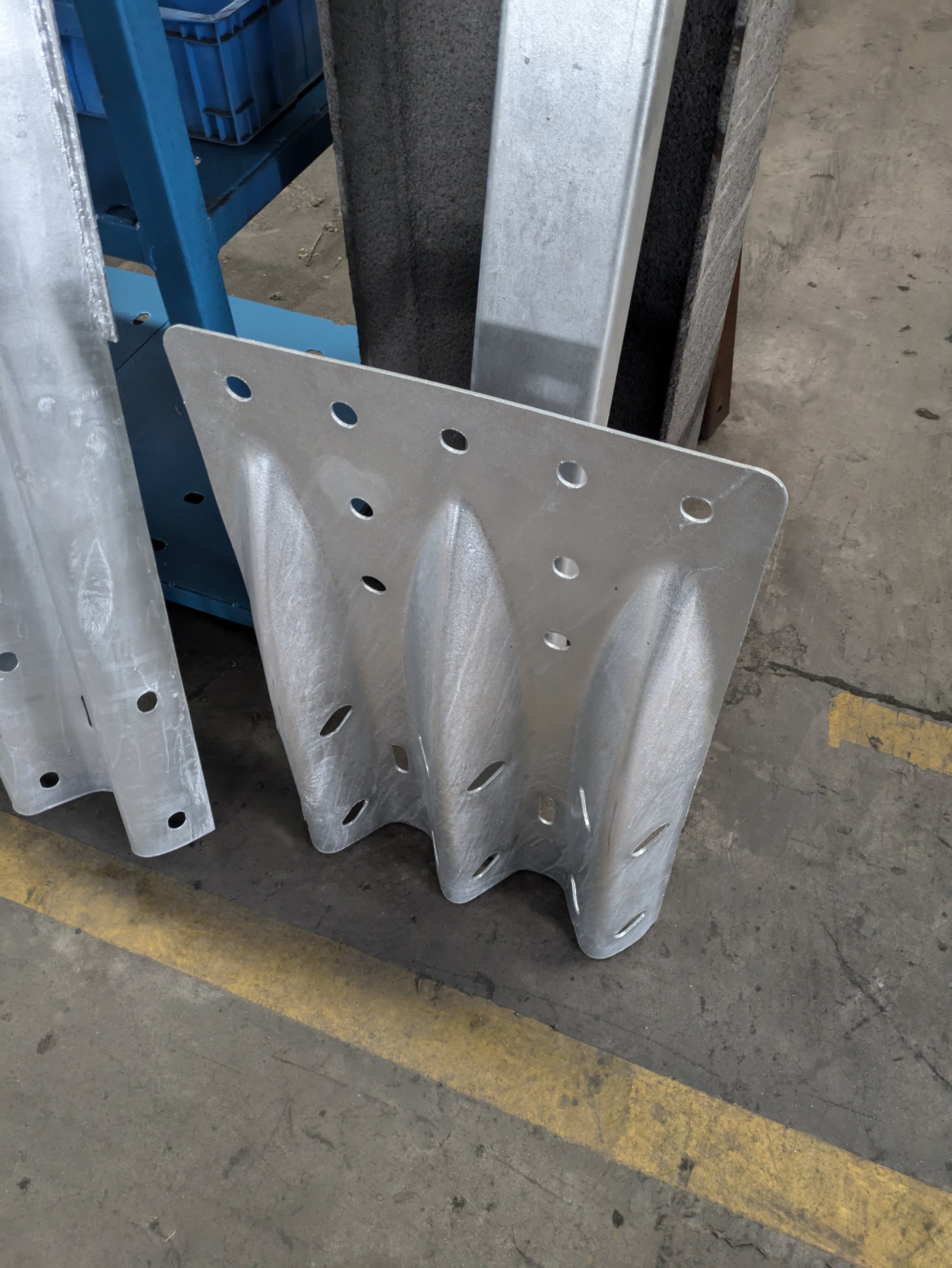 A metal bracket with multiple holes leaning against a stack of similar brackets in a warehouse or factory setting.