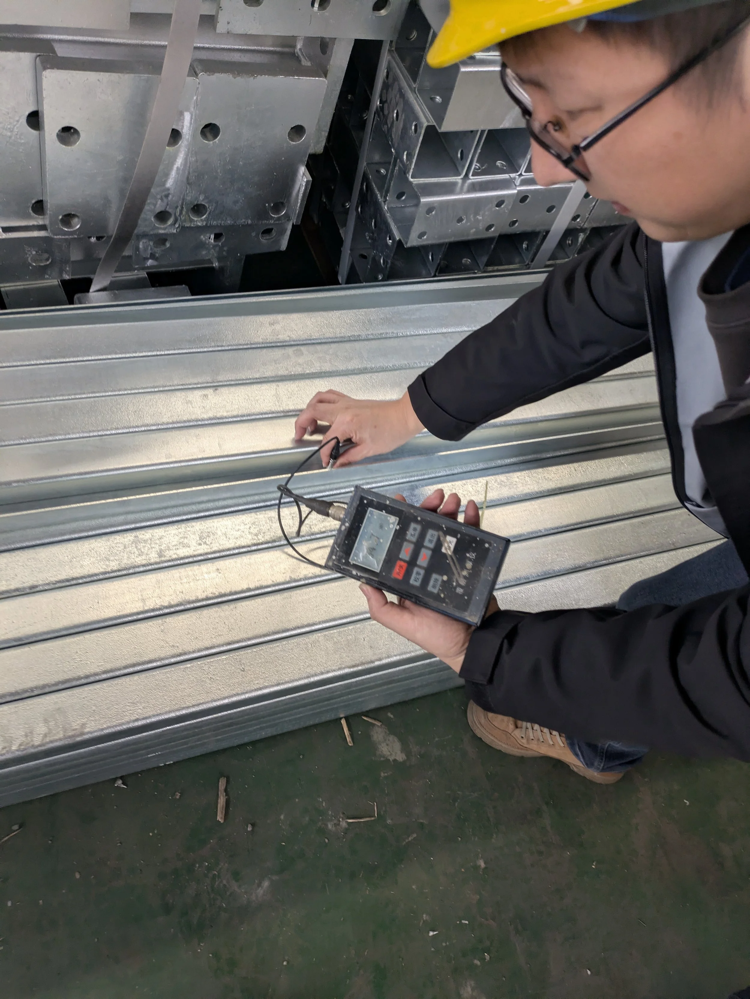 A person wearing a yellow hard hat and glasses is using a handheld device with a digital display and probes to inspect metallic metal roofing panels in an industrial setting.