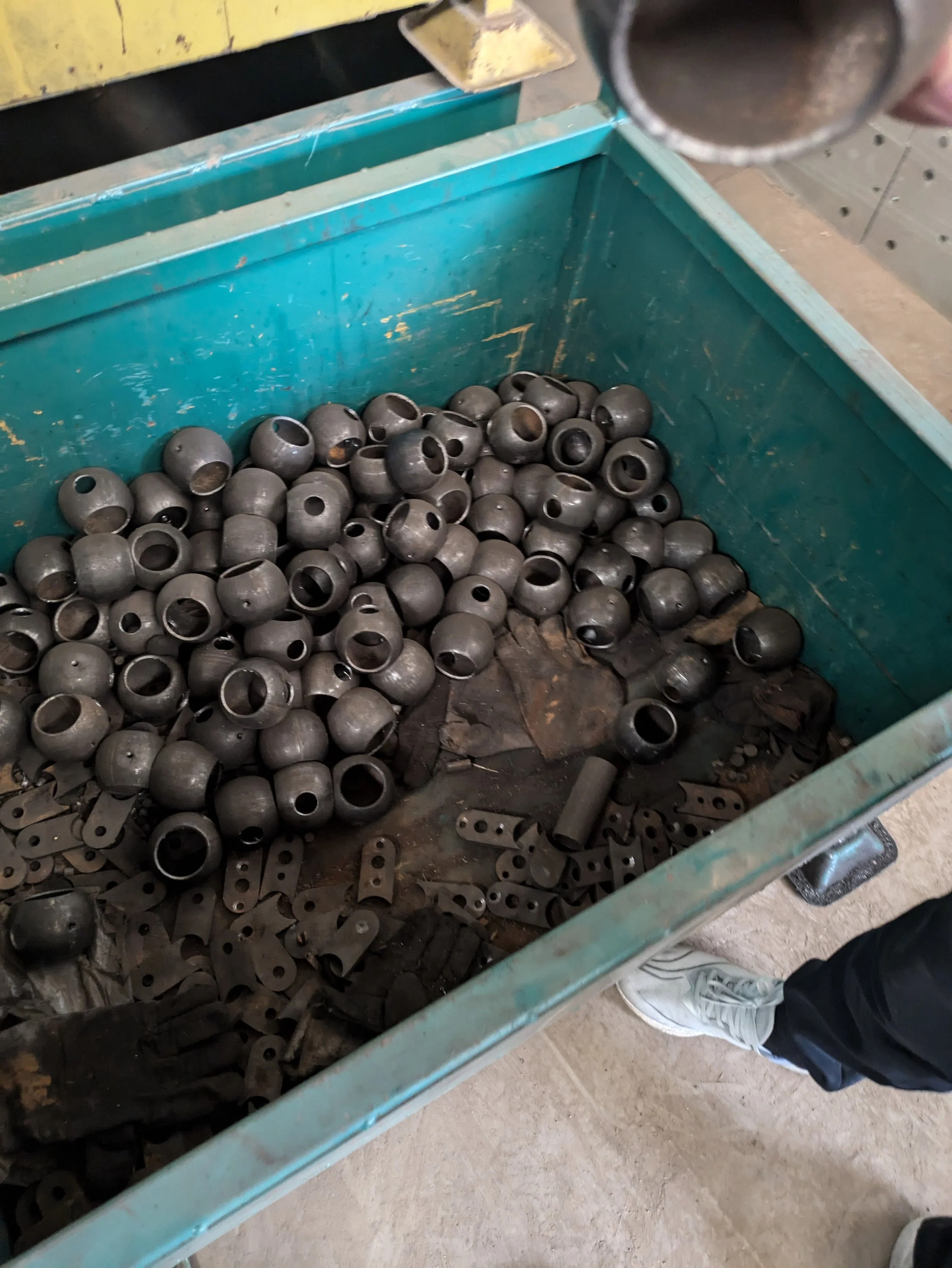 A green metal bin filled with numerous small black metal balls and scattered metal parts, with a person wearing a white shoe and black pants standing nearby.