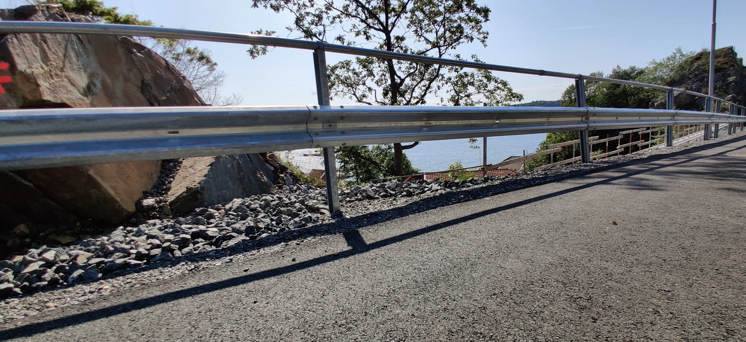 A paved road with a metal guardrail on the left side, rocks and a slope beside the road, trees, and a body of water in the background under a clear sky.