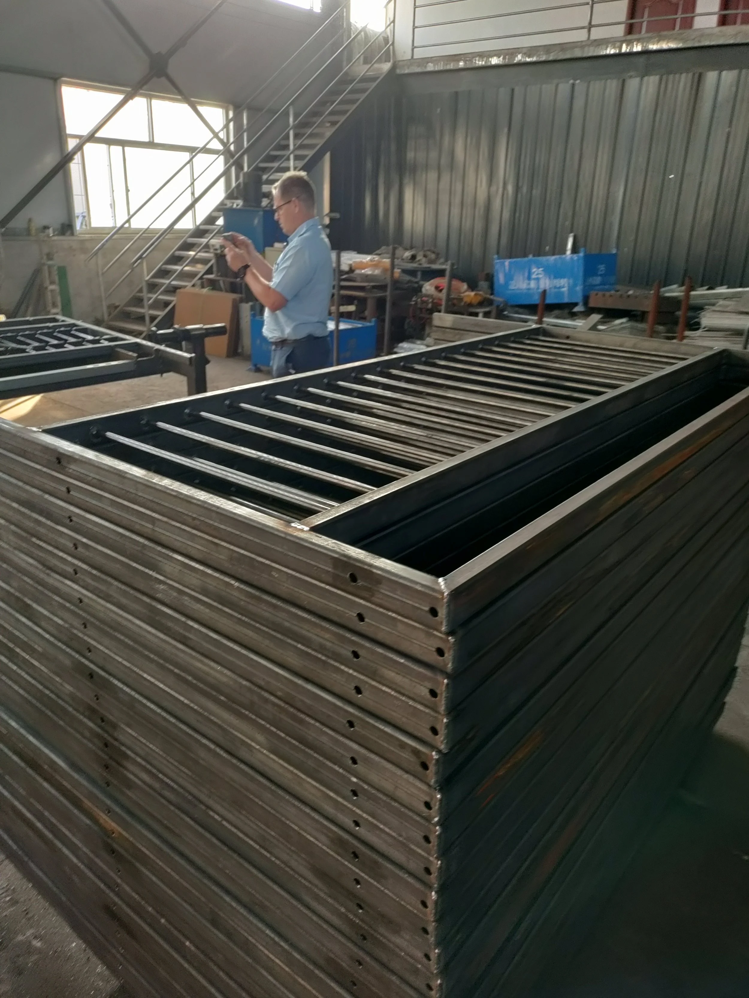 Stack of metal frames in an industrial workshop with a man in safety glasses and a light blue shirt standing nearby, using a smartphone.