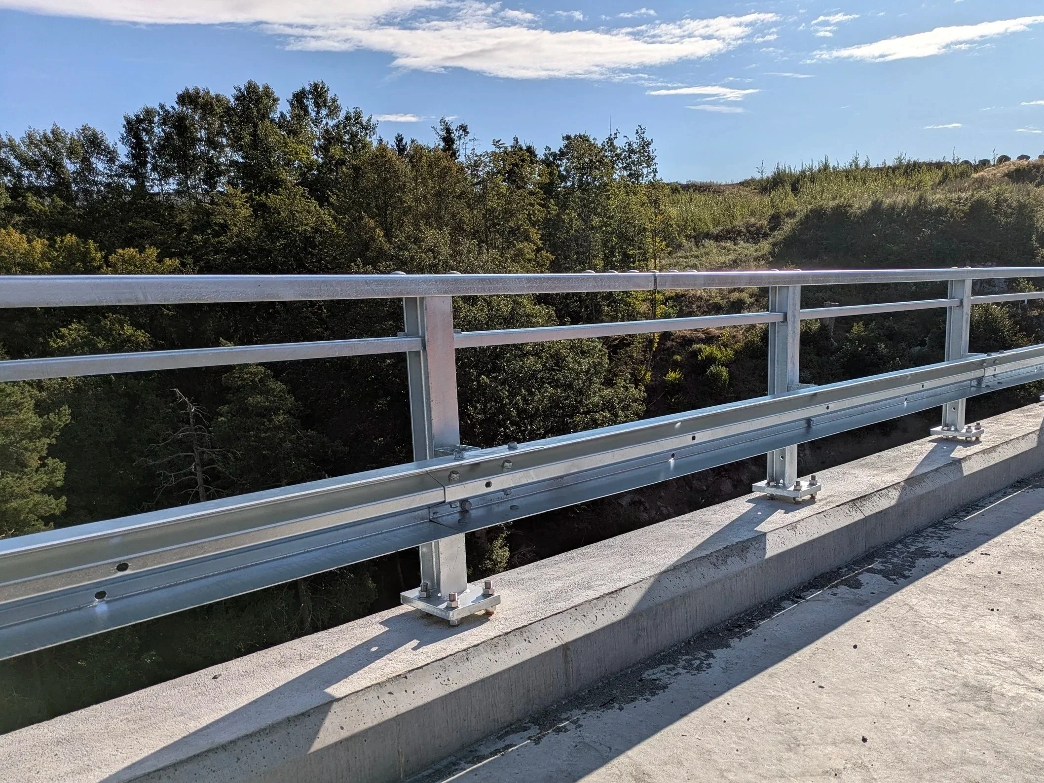 Metal guardrail along the edge of a concrete road with a view of trees and a hillside under a partly cloudy sky.