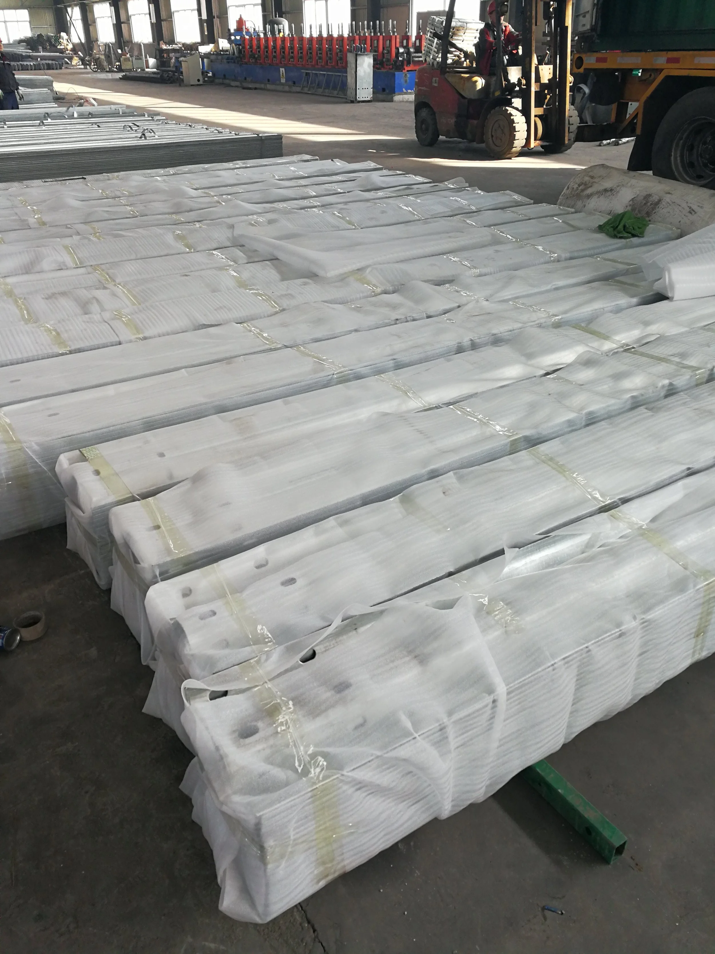 Stacks of white, wrapped, possibly metal or plastic, parts on a factory floor, with industrial equipment and trucks in the background.