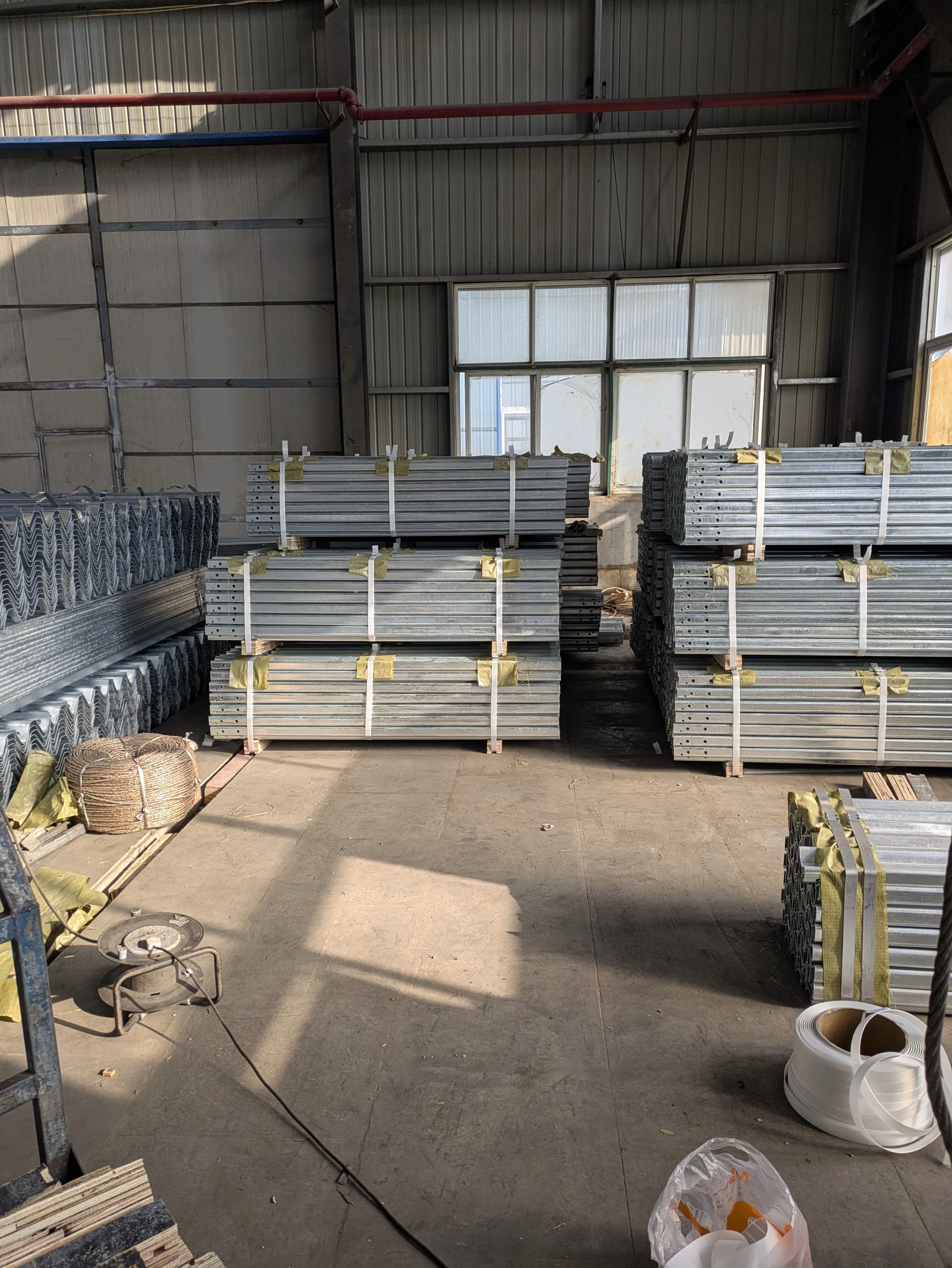 Stacks of metal pipes and fittings stored in a warehouse with sunlight streaming through windows.