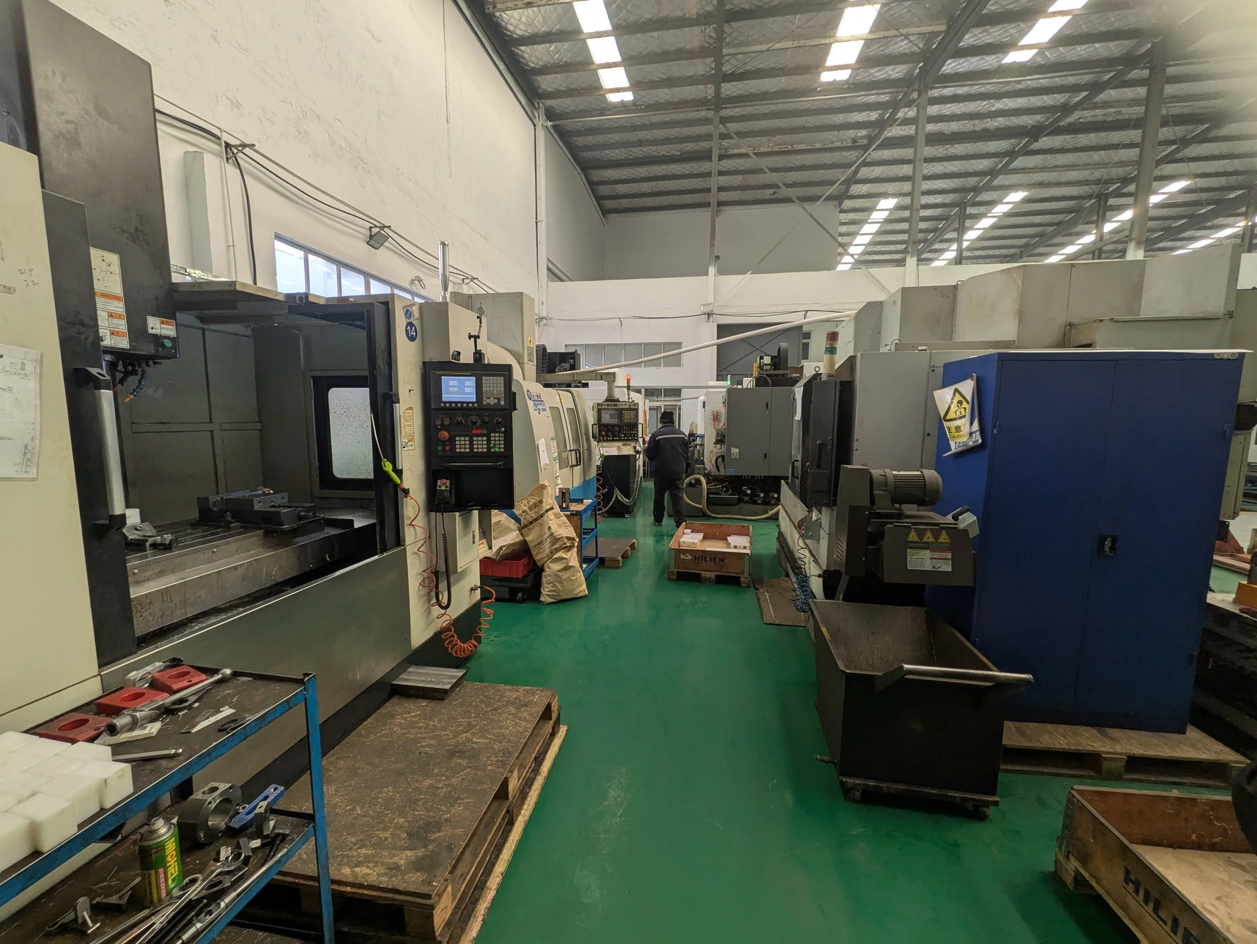 Industrial manufacturing shop with multiple CNC machines and tools, person working in the background, green polished concrete floor, high ceiling with metal roof, and large windows allowing natural light.