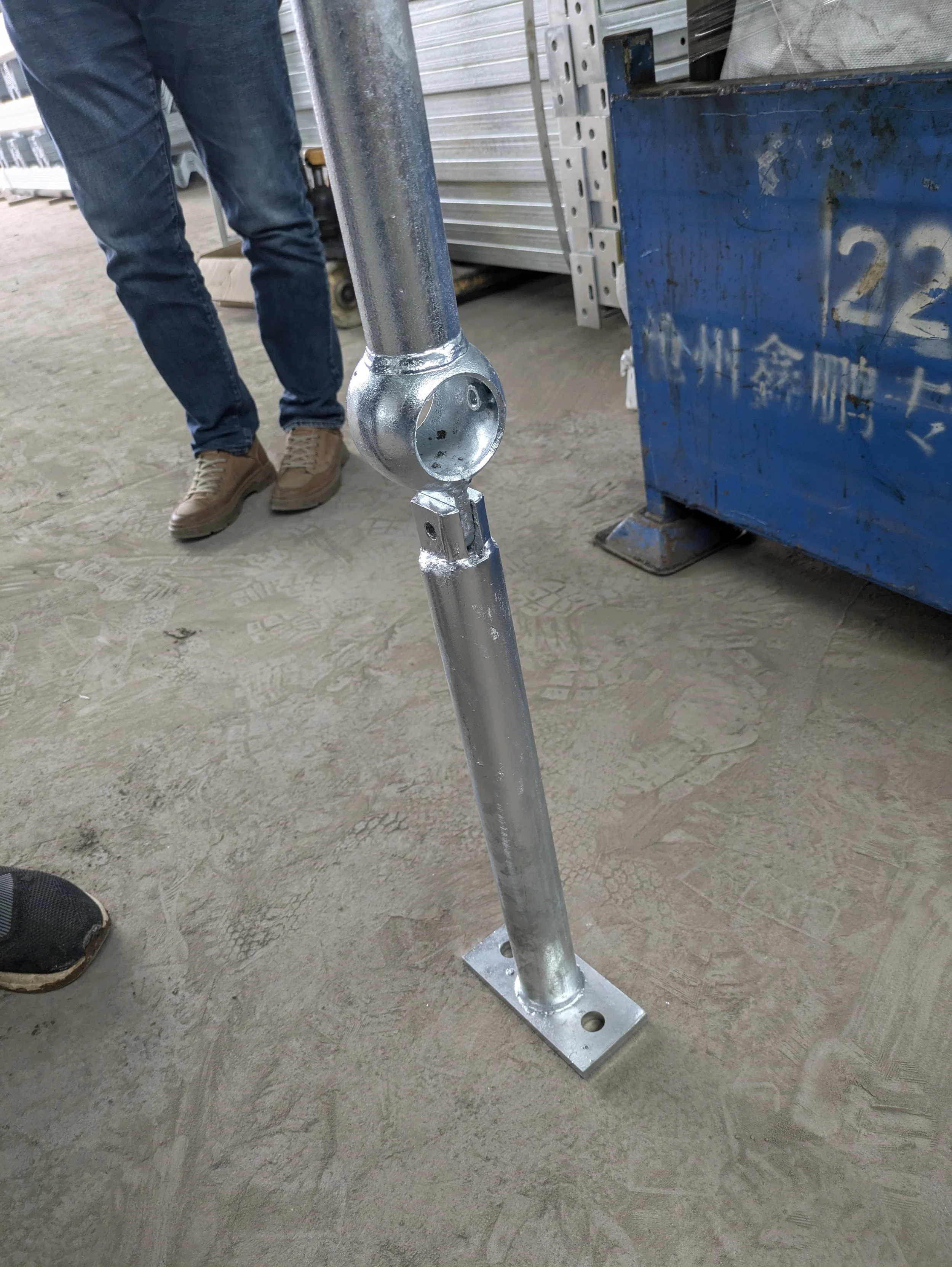 Metal scaffolding support pole with a round joint connector in a construction area, with workers nearby and construction materials in the background.