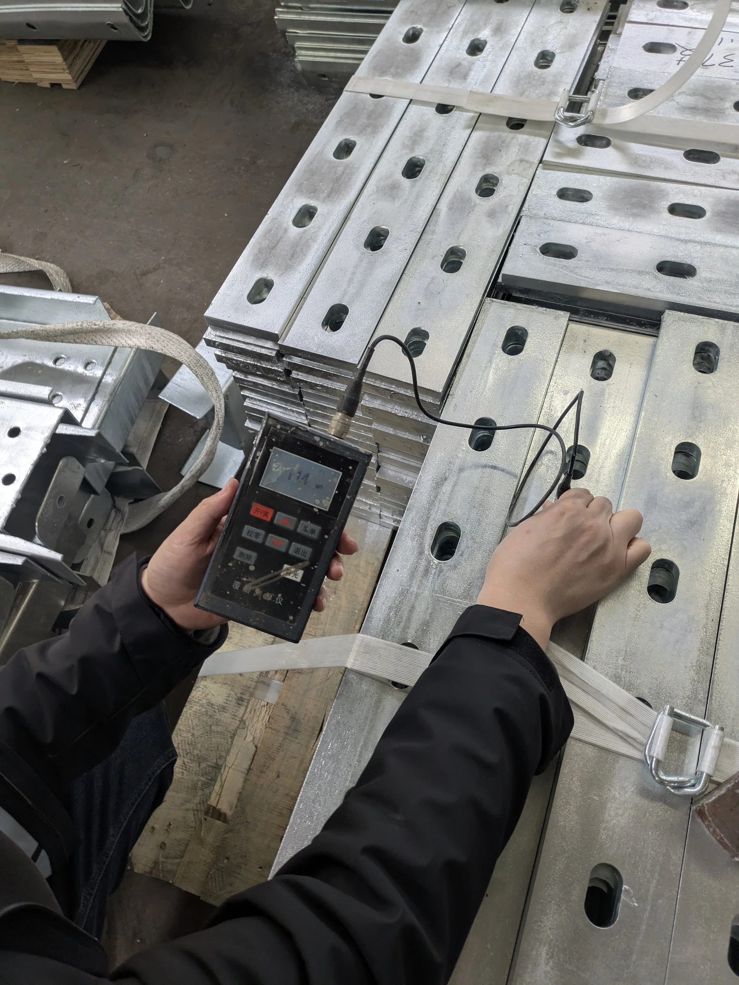 Person measuring the thickness of metal plates stacked on a wooden surface with a digital thickness gauge in a workshop or manufacturing setting.