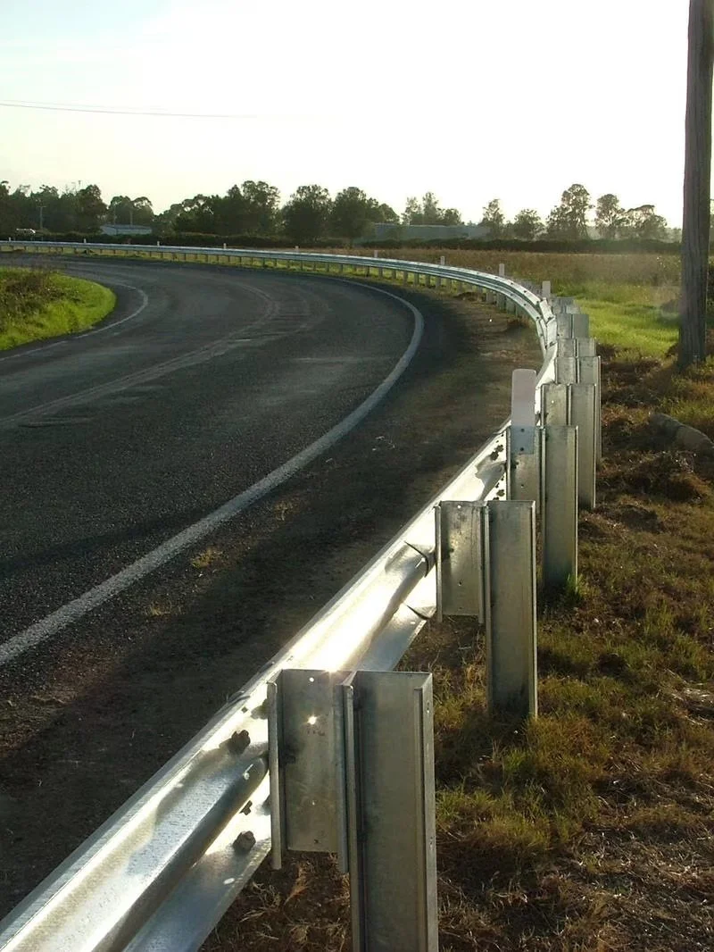 A curved rural road with a metal guardrail on the right side, green grass along the roadside, and trees in the background under a clear sky.
