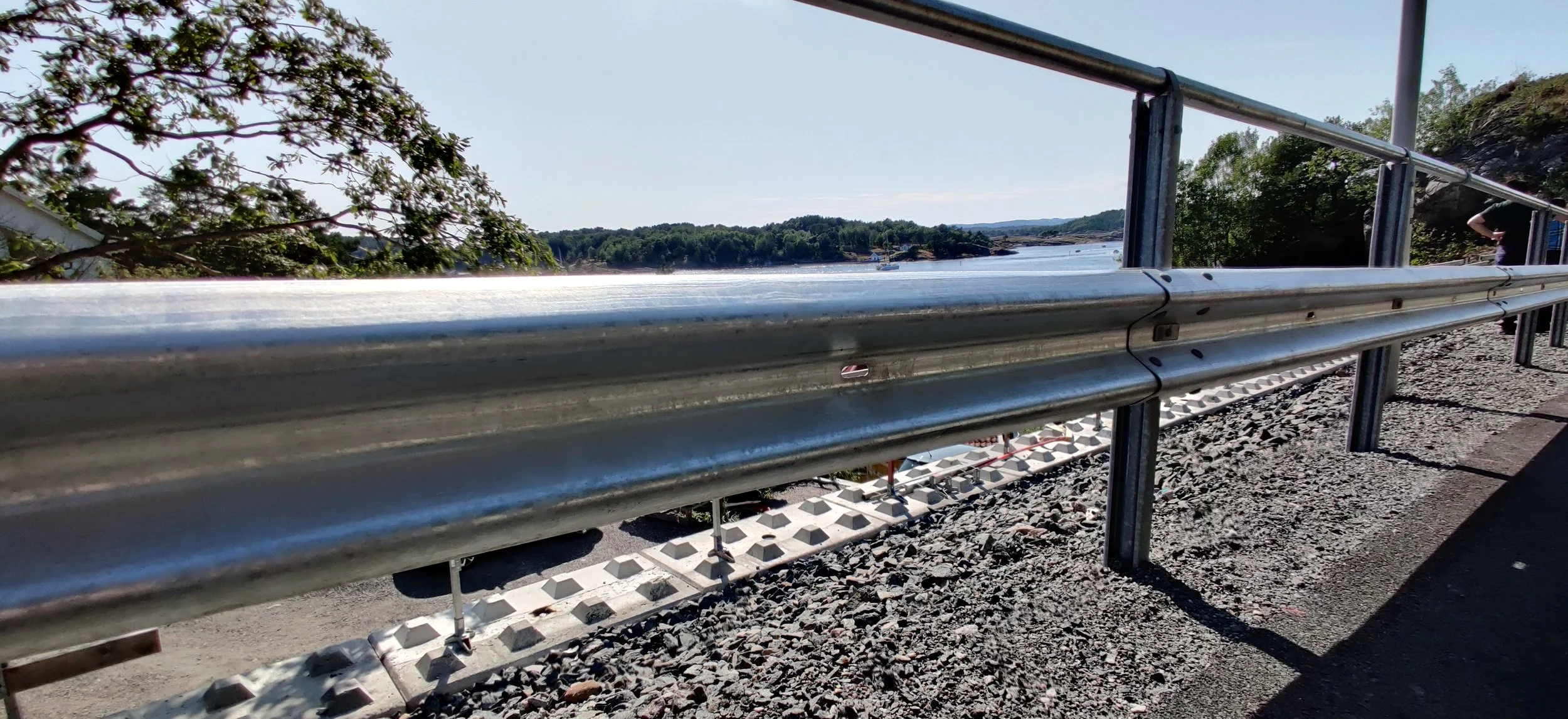 Metal guardrail along a road overlooking a body of water with trees and hills in the background, under clear blue sky.