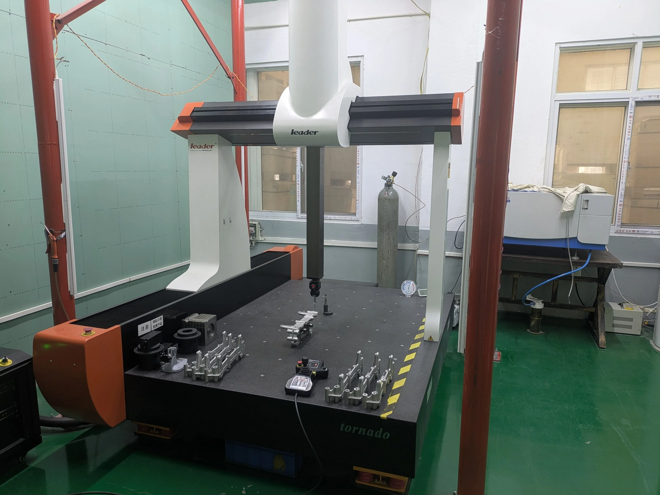 A large industrial machine with a curved white arm labeled 'leader' and a black horizontal bar, situated in a room with green flooring, red structural poles, and windows in the background. Various tools and metal components are arranged on the black 