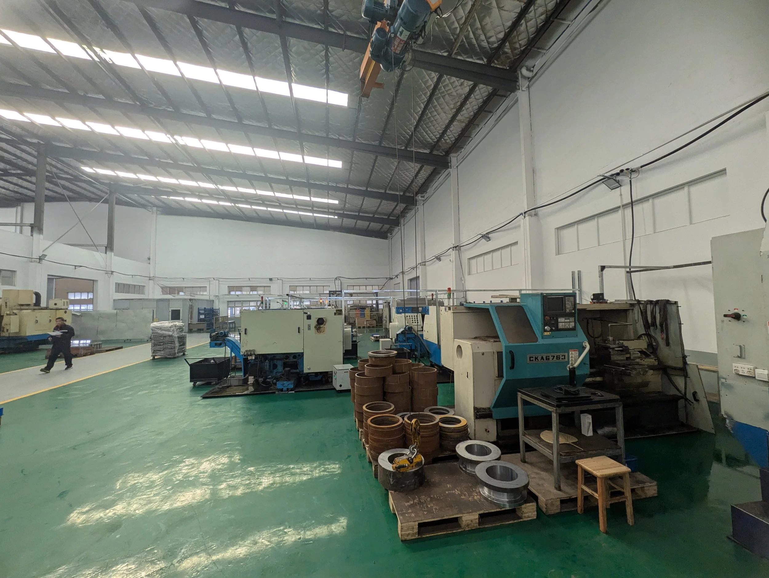 Inside an industrial manufacturing facility with various machines and equipment, including a row of CNC lathes, stacks of metal parts, and a worker walking along the green polished floor under a high ceiling with skylights.
