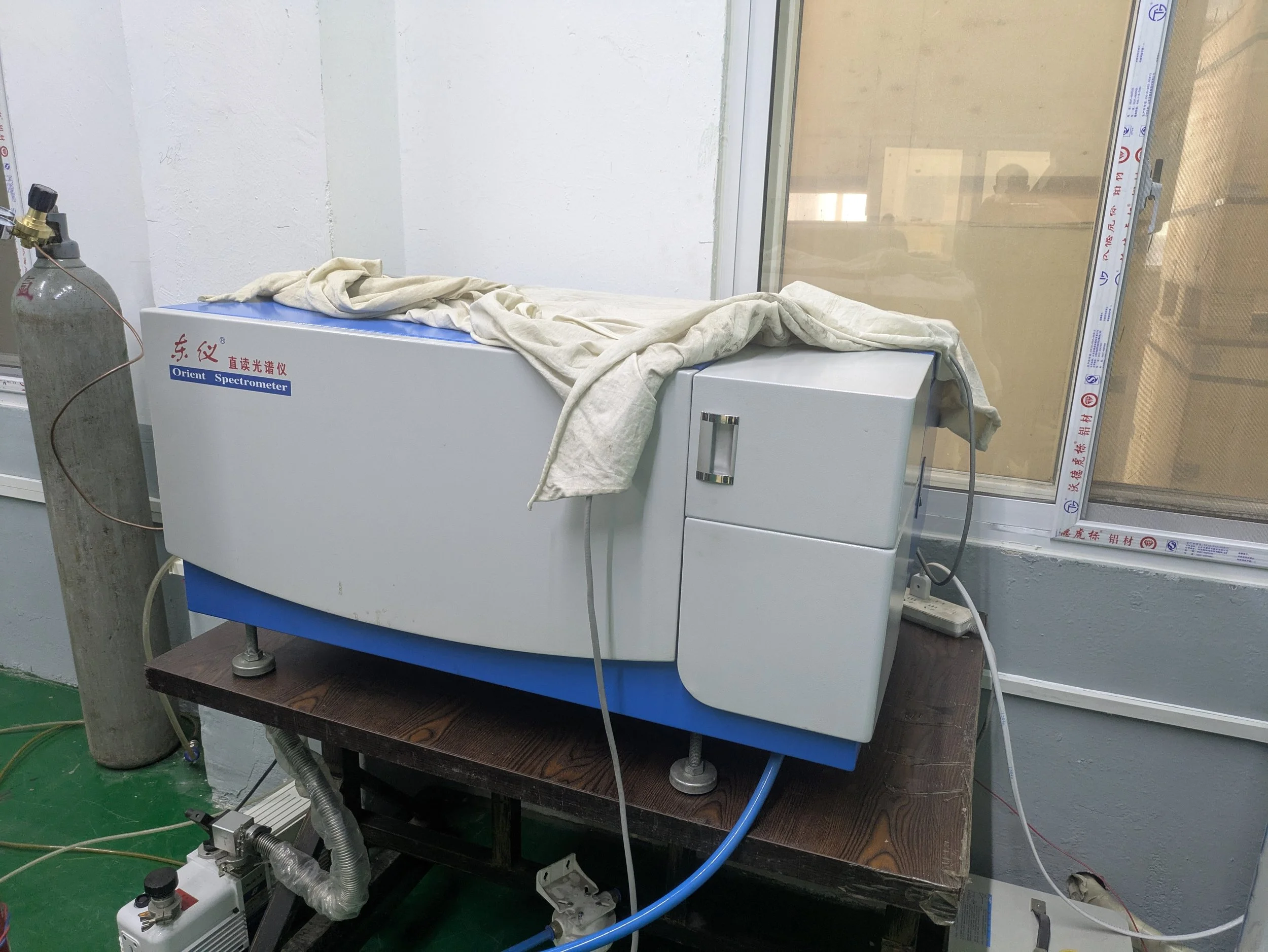 Laboratory spectrometer machine with a wet cloth draped over it, placed on a wooden table next to a window, in a scientific lab setting.
