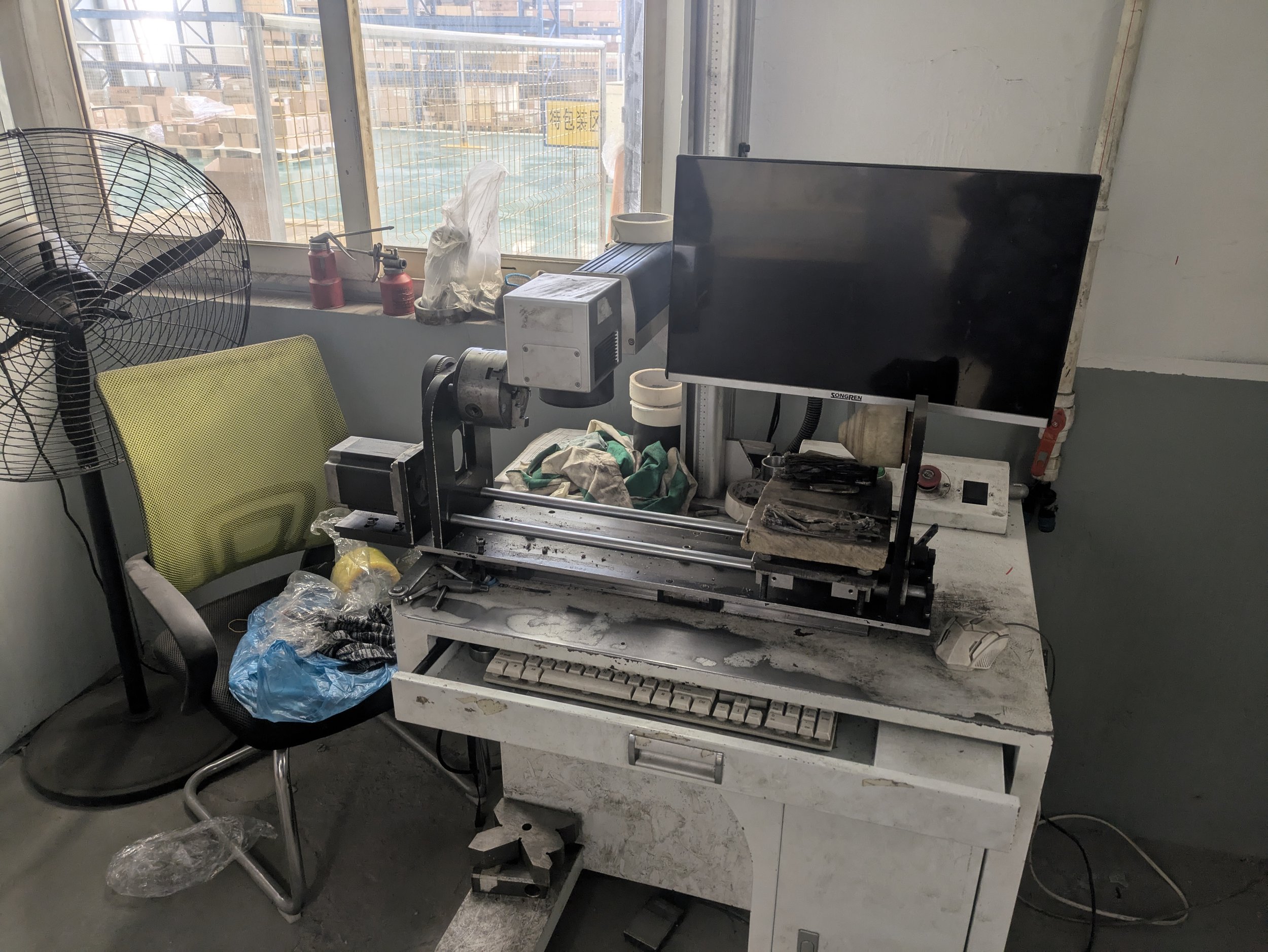 Dirty industrial workspace with a modern machine on a cluttered white desk, a computer monitor, and a green office chair, next to a standing fan, in a room with a large window showing a construction site outside.