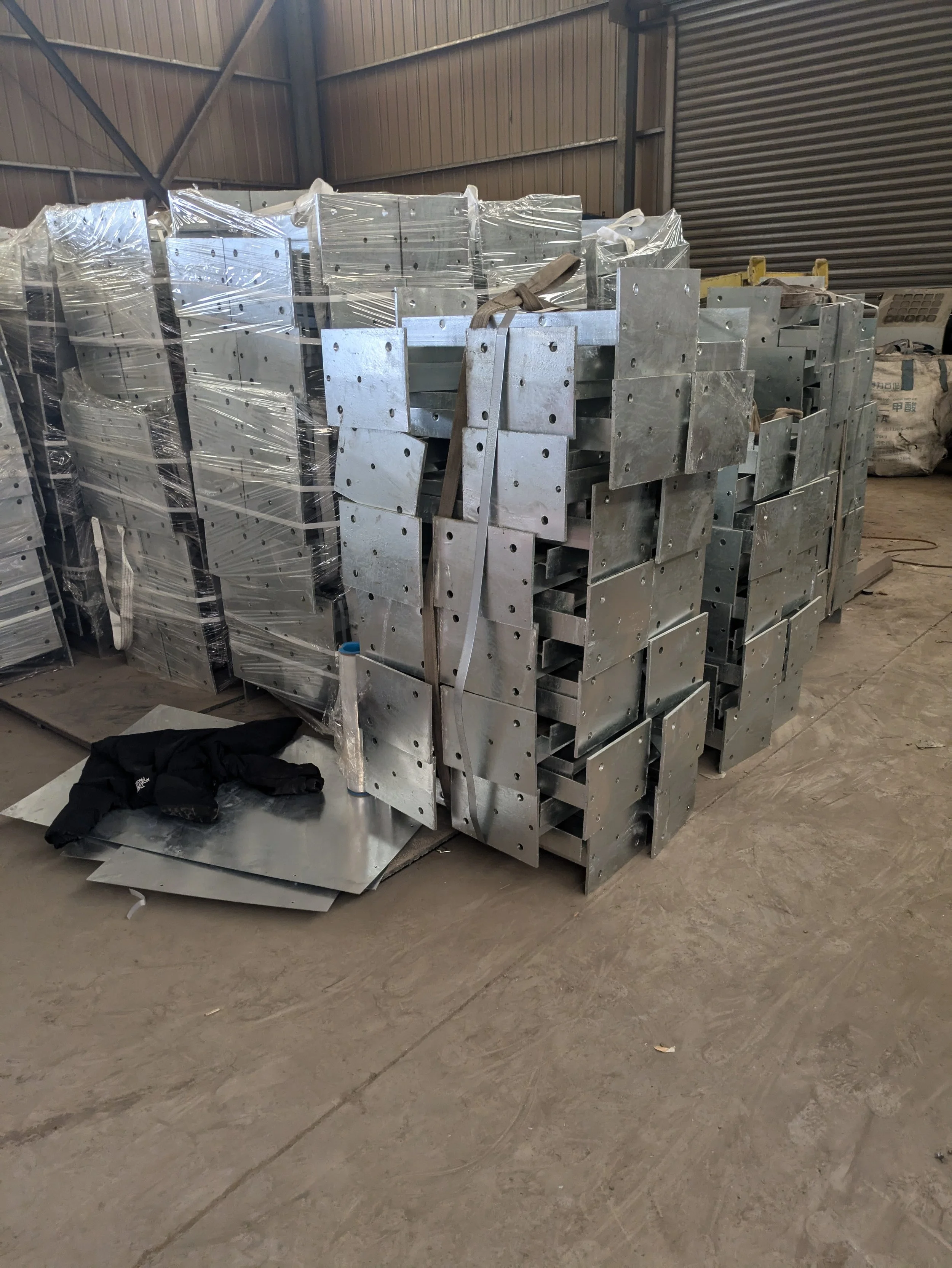 Stacks of metal parts and panels in a warehouse, some wrapped in plastic, with a pair of black gloves on a sheet of metal on the ground.
