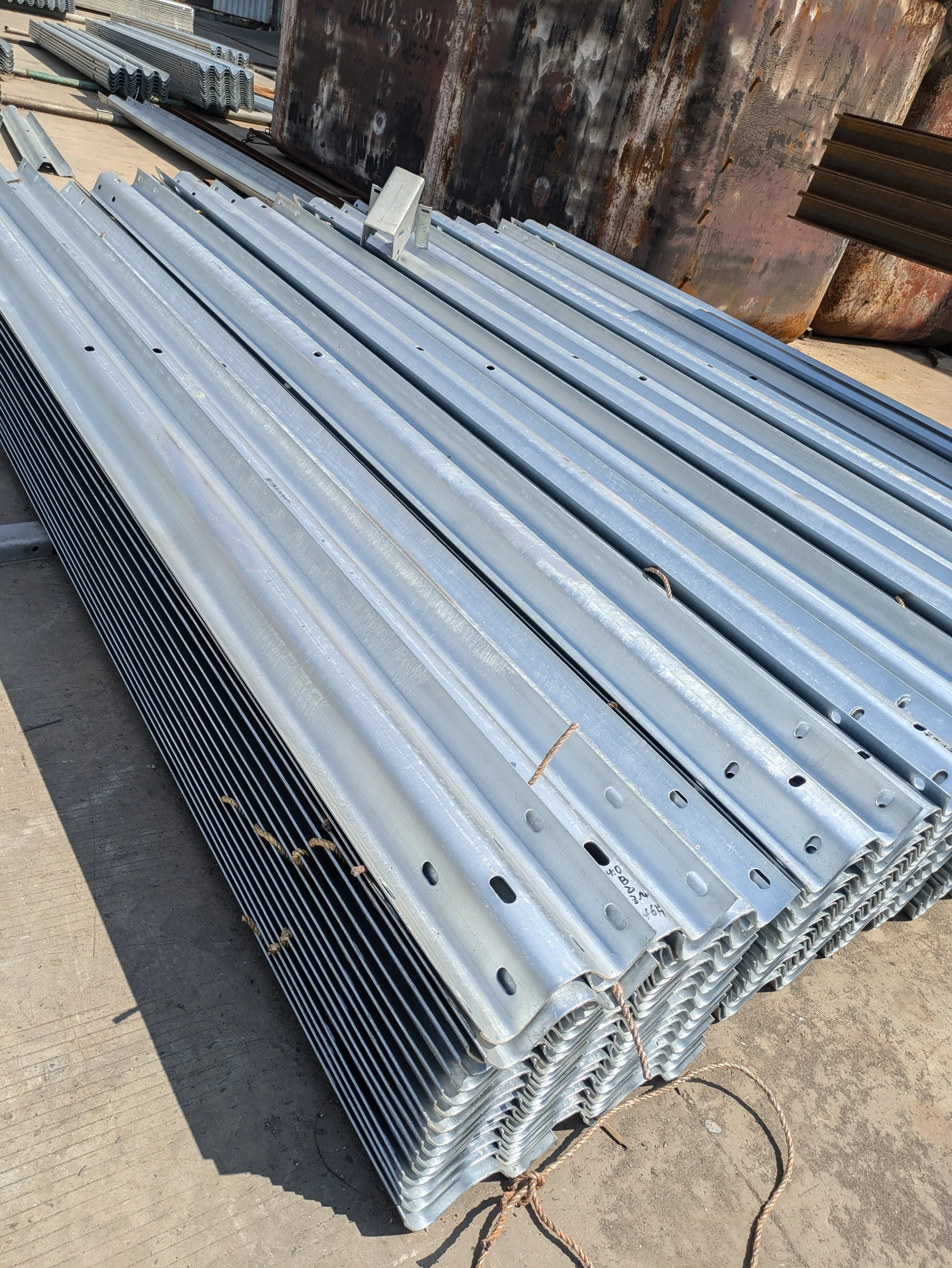 Stacked metal beams with holes and ridges, tied together with brown rope, outdoors on concrete ground, rusted large tanks in background.