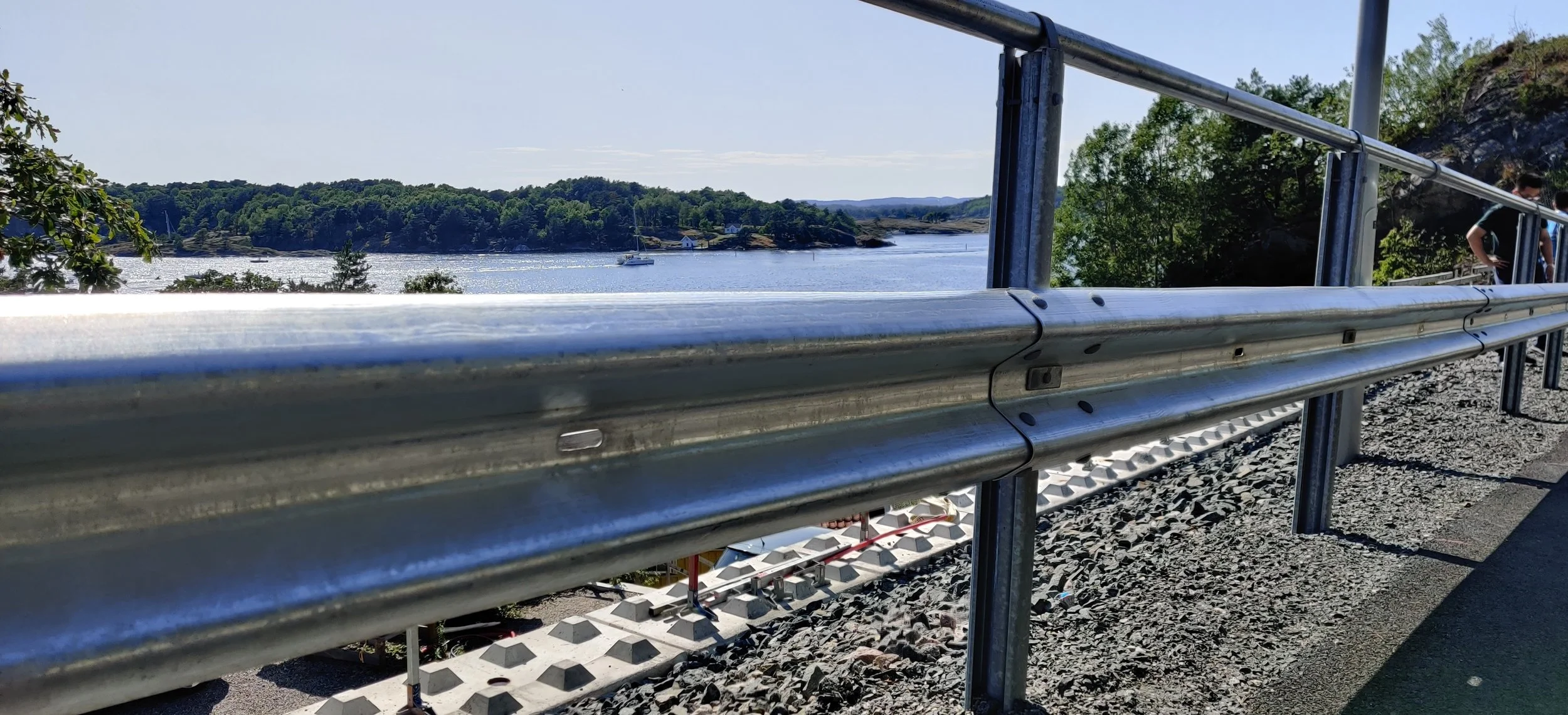 Close-up of a metal guardrail along a scenic lakeside road with trees, water, and a boat in the background on a sunny day.