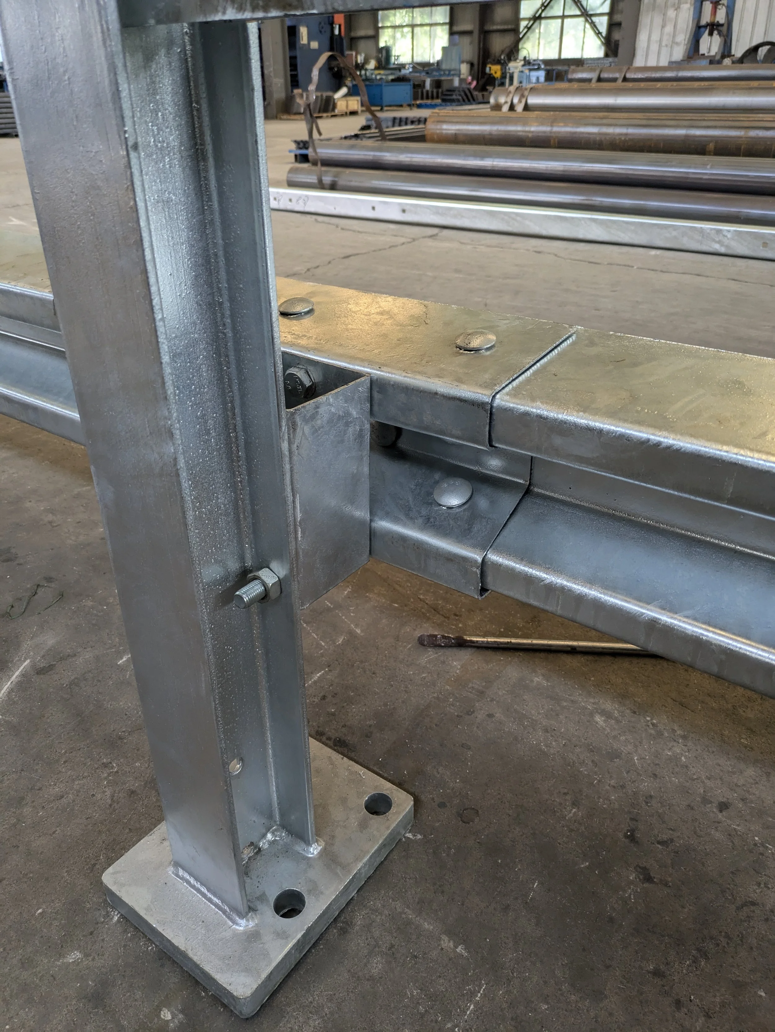 Close-up of a metal frame and bolted joint in an industrial or workshop setting, with steel pipes and equipment in the background.