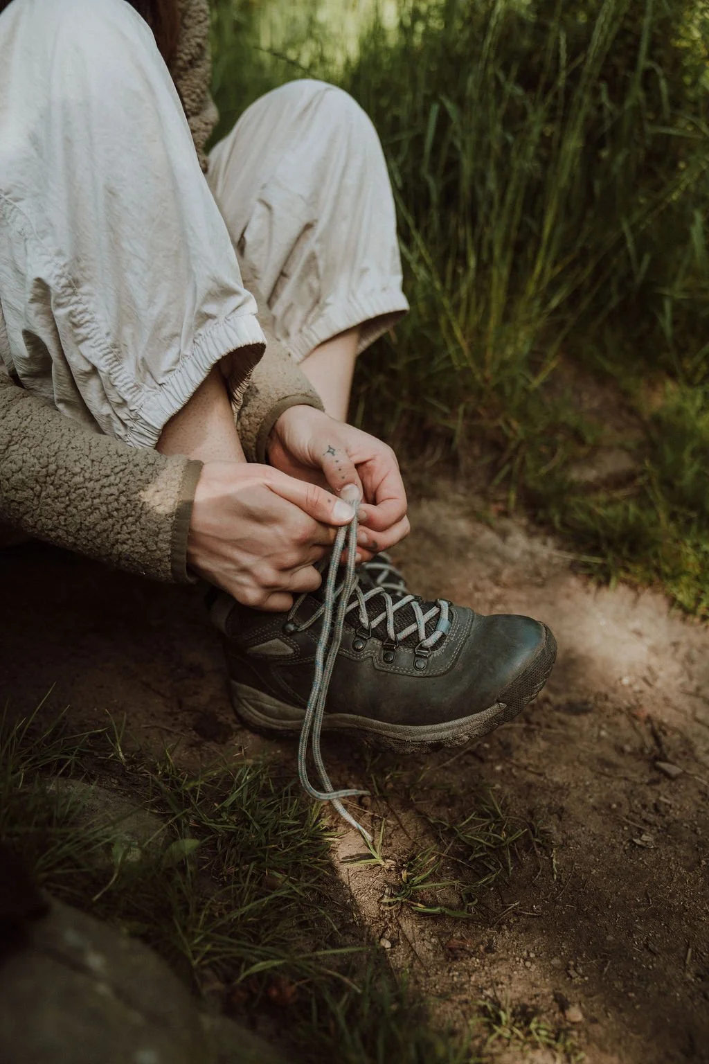A person wearing hiking boots, khaki cargo shorts, and a fleece jacket, sitting on the ground and tying their shoelaces in a natural outdoor setting with grass and dirt.