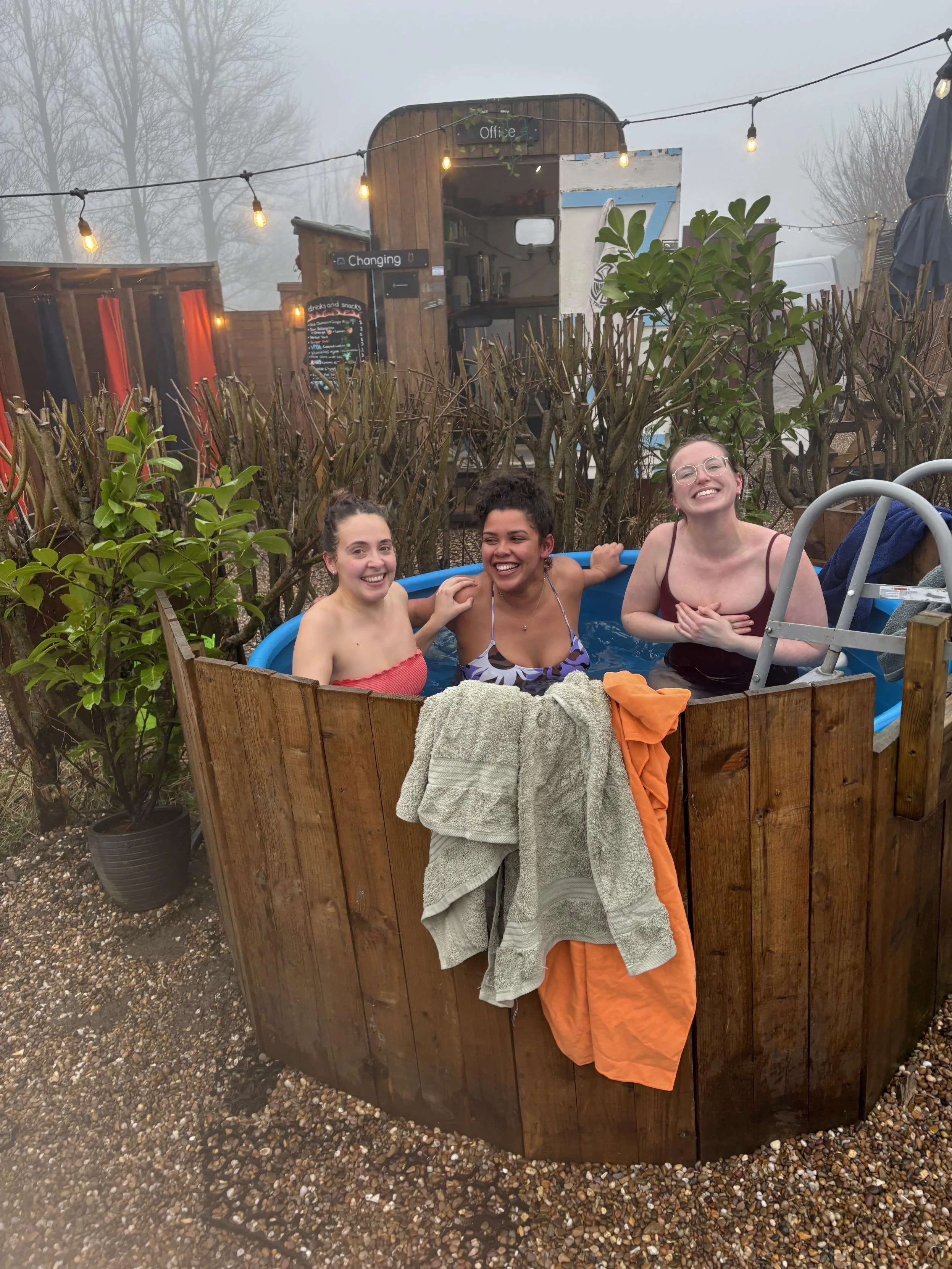 Three women smiling and laughing while sitting in a small blue plastic pool outdoors, surrounded by a wooden fence and plants, with a foggy background and outdoor string lights.