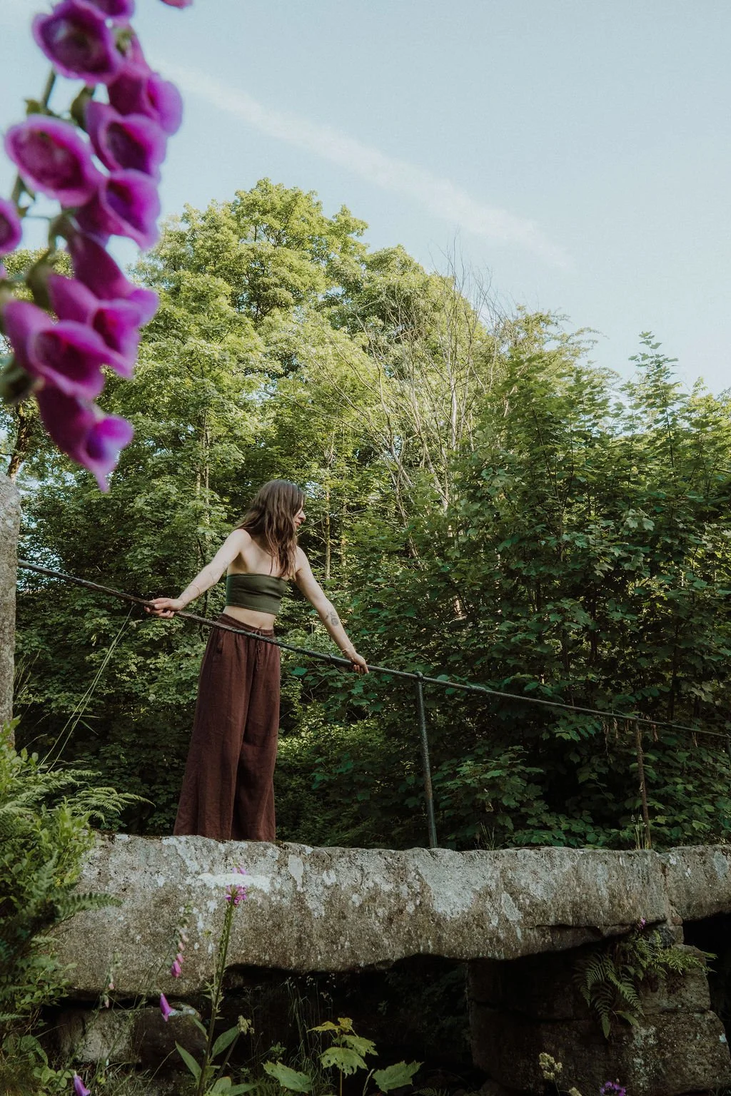 A woman with long hair, wearing a green crop top and wide brown pants, stands on a stone bridge holding onto a rope railing, surrounded by dense green trees and foliage.