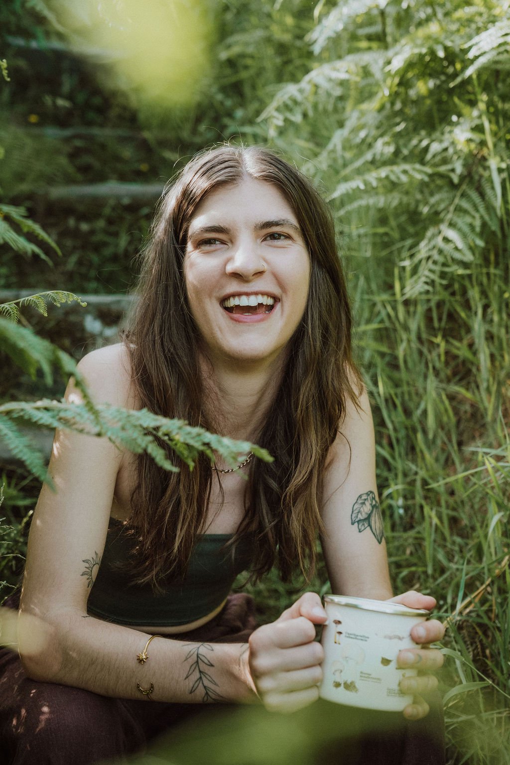 A young woman with long brown hair smiling while sitting in a lush, green forest, holding a white enamel mug, with tattoos on her arms and a necklace, enjoying nature.