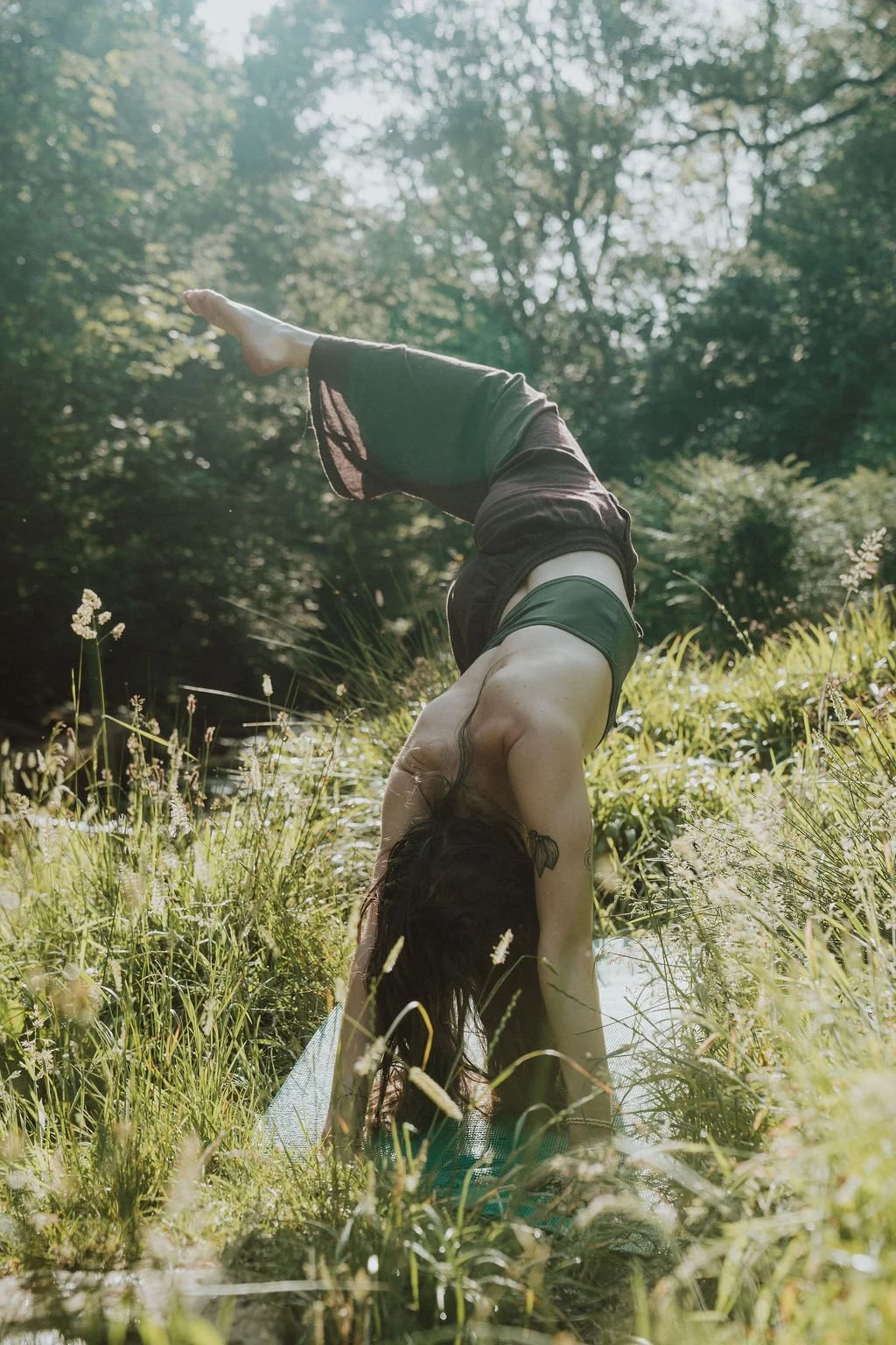A woman practicing yoga outdoors in a grassy area near a river, performing a backbend pose with greenery and trees in the background.