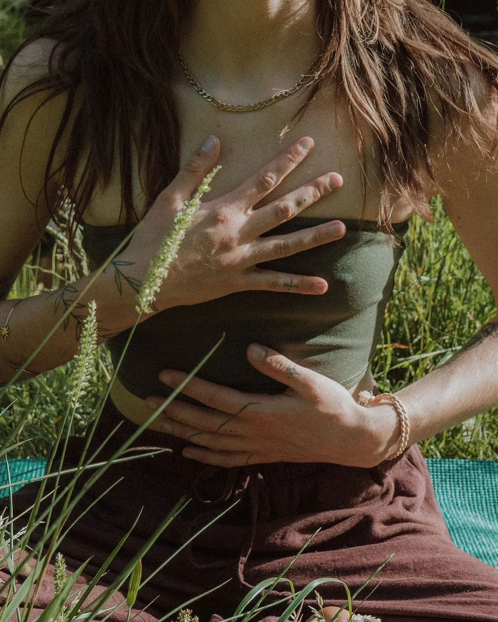 A person with long hair wearing a gold chain necklace and a dark green tube top is sitting outdoors amid green plants, holding their chest with one hand and their stomach with the other.
