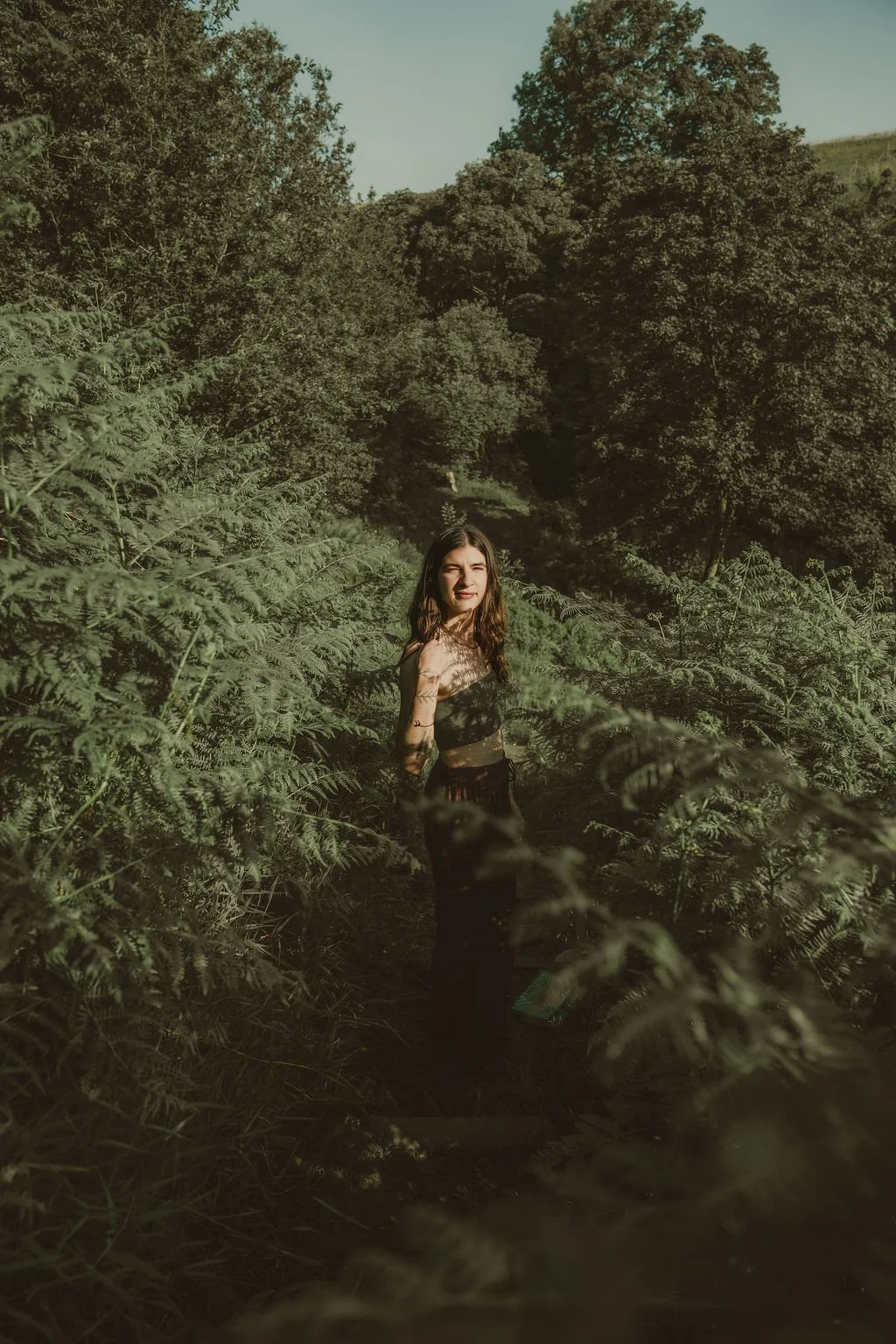 Woman standing among dense fern plants in a forested area with trees and hills in the background during daytime.