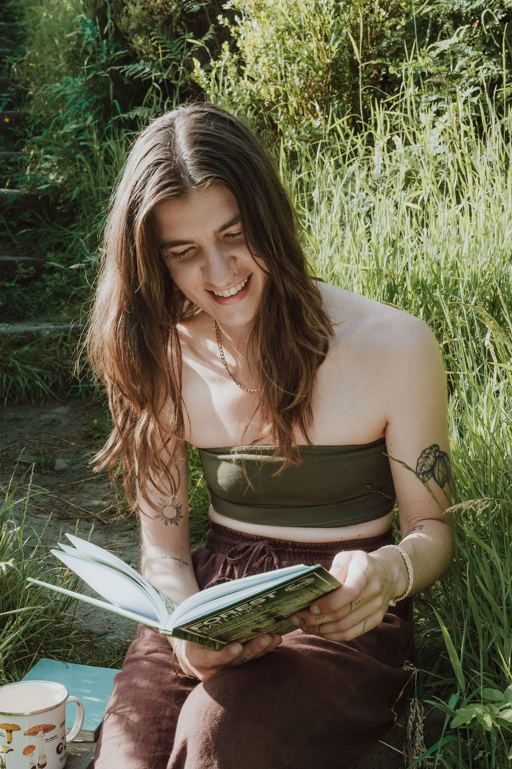 Young woman with long brown hair, tattoos, and a nose ring, sitting outdoors in a grassy area, reading a book, smiling, wearing a green strapless top and brown pants.
