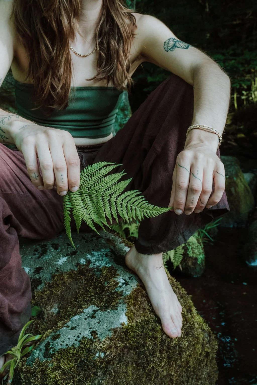 Person sitting on mossy rocks holding a fern leaf, with long hair, tattoos on arms, wearing a dark green tube top and brown pants, in a lush, green outdoor setting.