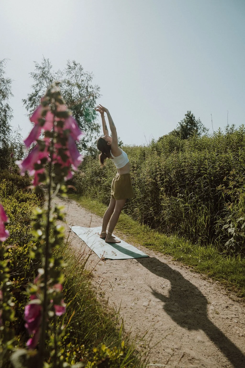A woman practicing yoga outdoors on a dirt path, surrounded by green plants and trees, with pink flowers in the foreground and the sun shining down.