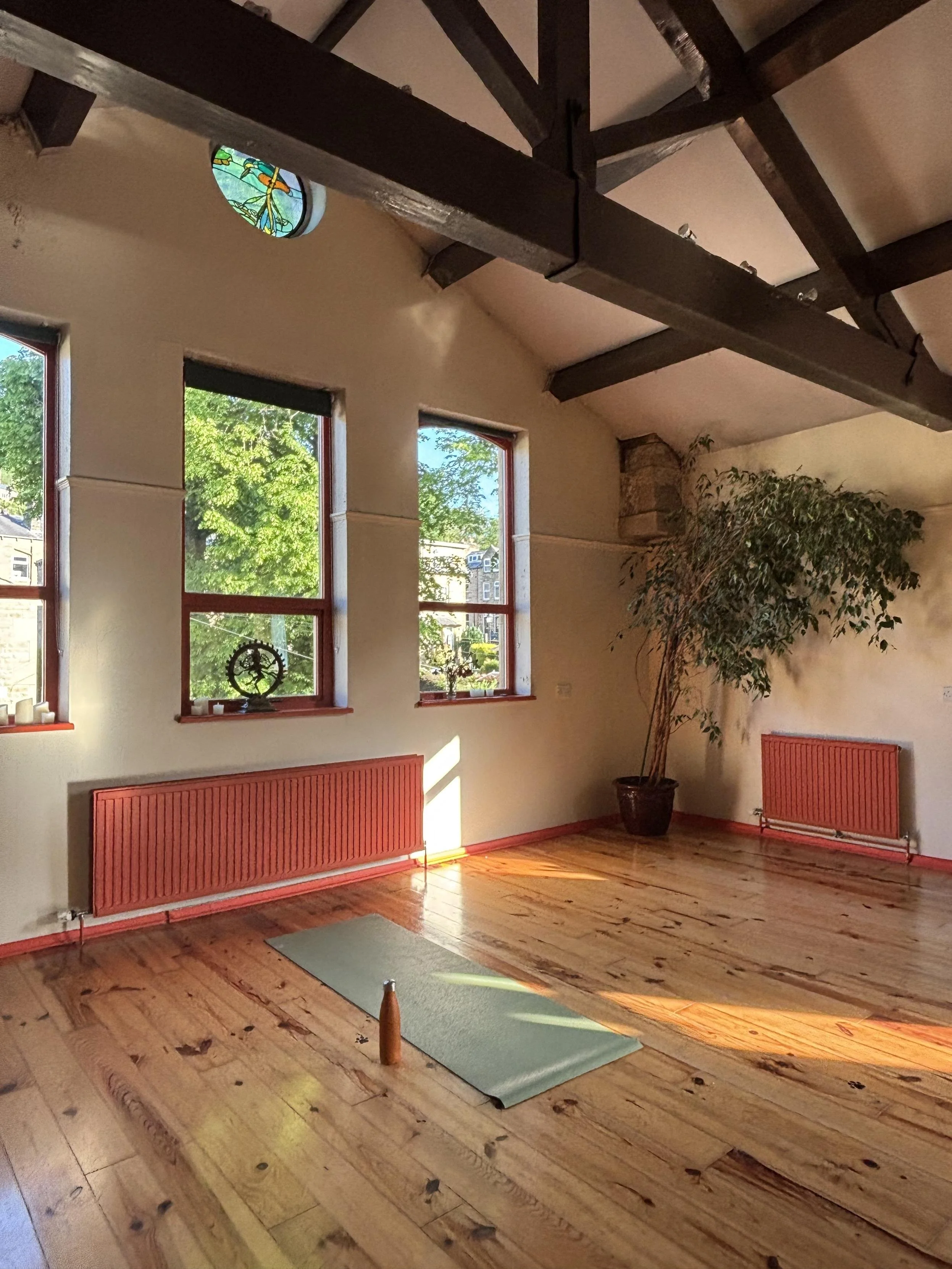 A room with wooden floors, four windows showing green trees outside, and a potted plant in the corner. There is a gray yoga mat on the floor with a small orange cone at the top of it. Two red radiators are mounted on the wall beneath the windows, and some decorative items are placed on the windowsills.