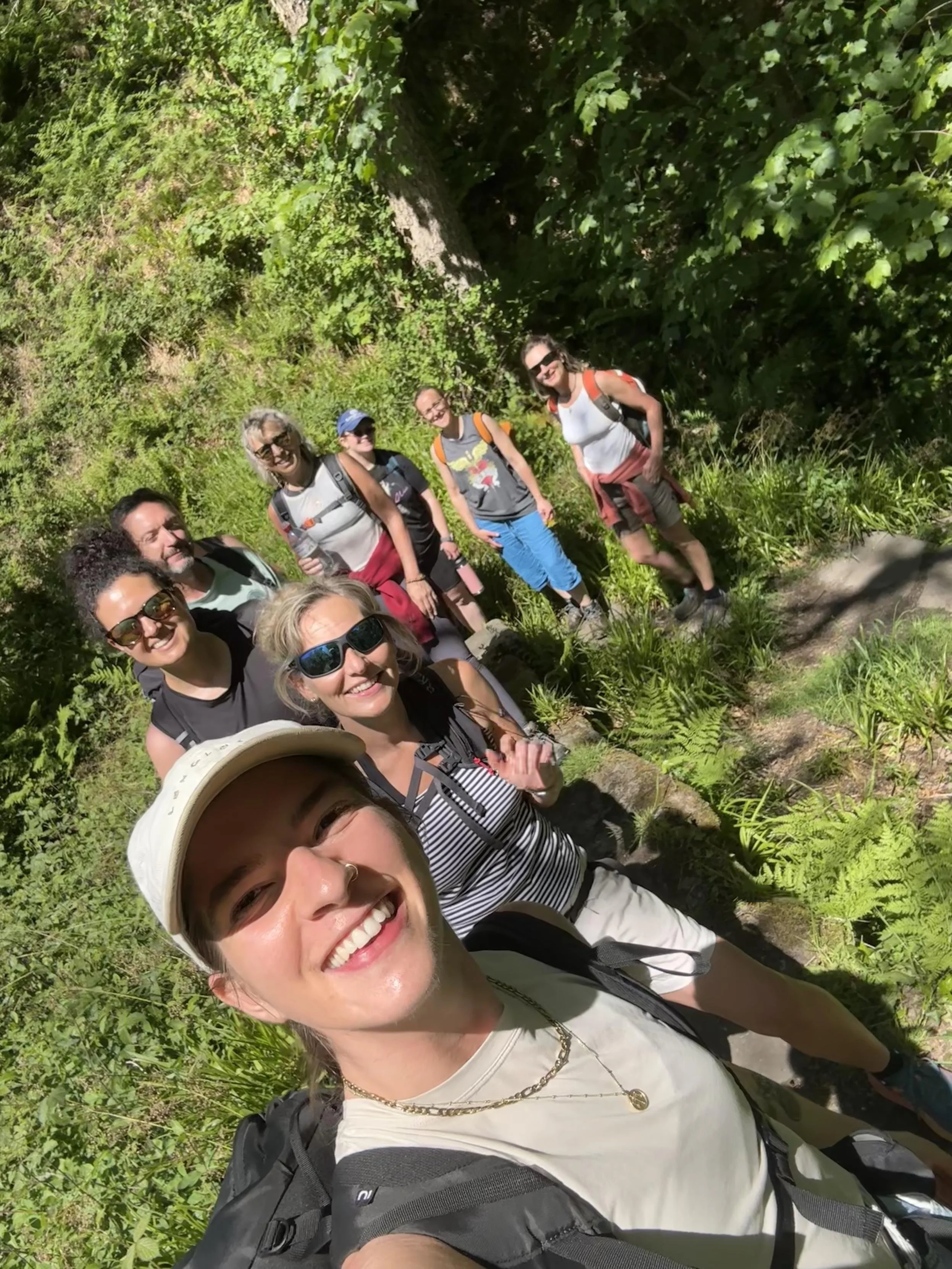 A group of smiling hikers taking a selfie outdoors in a green, wooded area with tall grass and trees.
