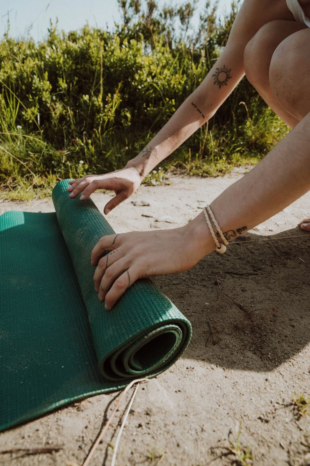 Person rolling out a green yoga mat outdoors on sandy ground near green bushes in sunlight.