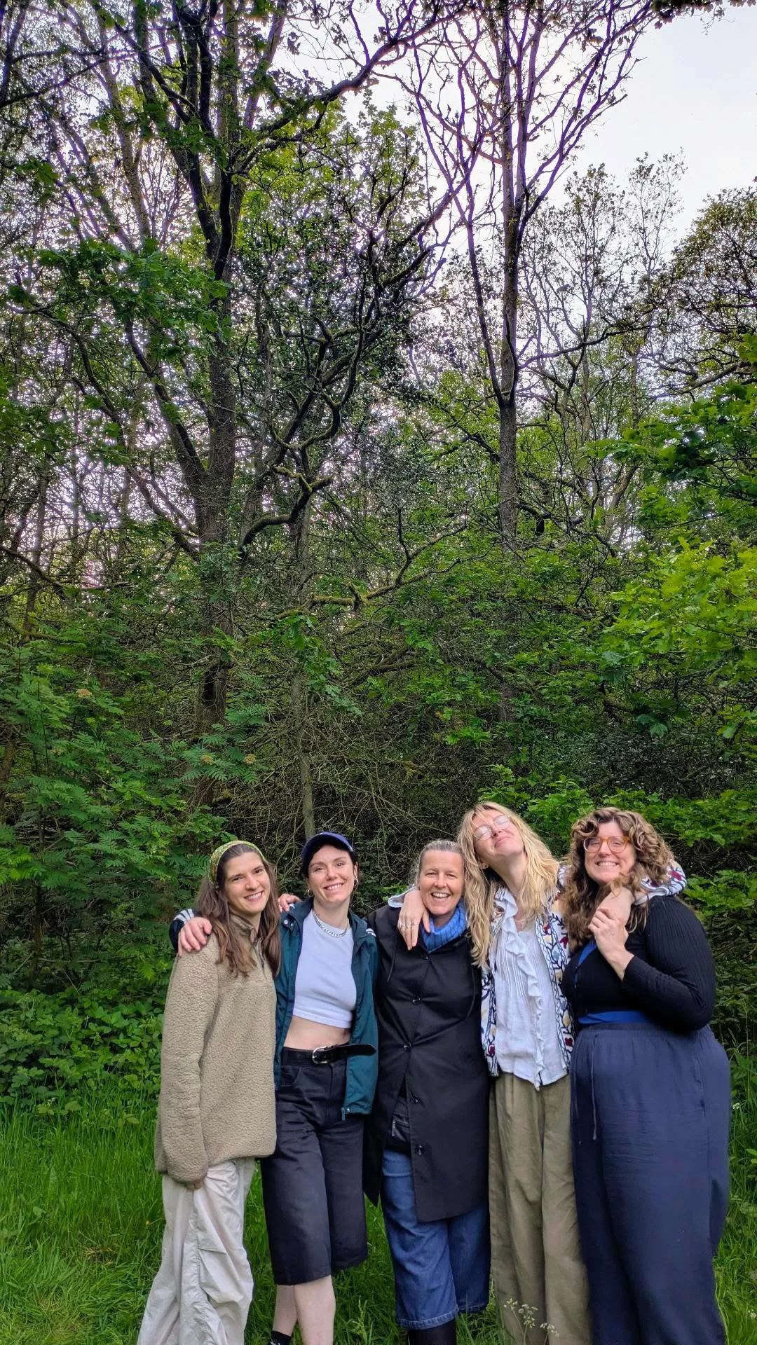 A group of five women standing outdoors in a forested area, smiling and embracing each other, surrounded by lush green trees and grass.