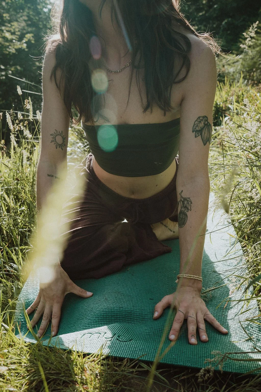A woman practicing yoga outdoors on a green mat in a grassy area, wearing a strapless black top and dark pants, with tattoos on her arms and a necklace.