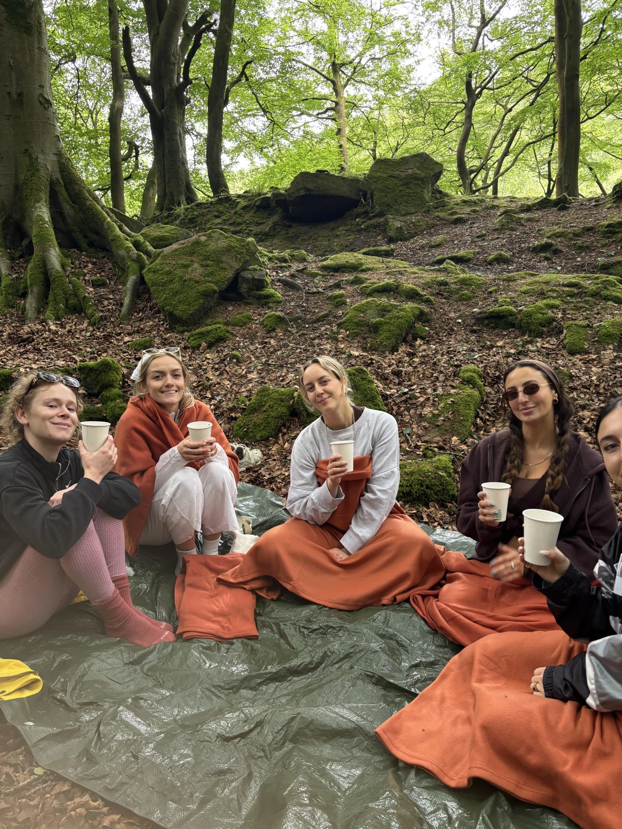 Group of five women sitting on a large tarp-covered spot in a forest, holding cups, with moss-covered rocks and green trees in the background.