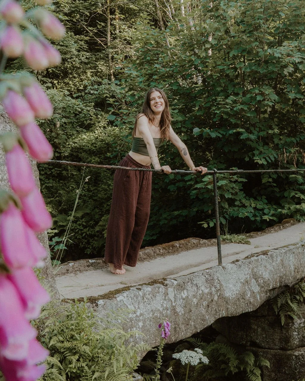 A woman with long hair smiling and holding a metal railing on a stone bridge in a lush forest, with pink flowers in the foreground.